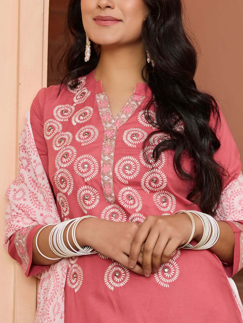 A woman wearing a pink embroidered rayon suit set with a kurta, pants, and a dupatta, standing in a doorway.