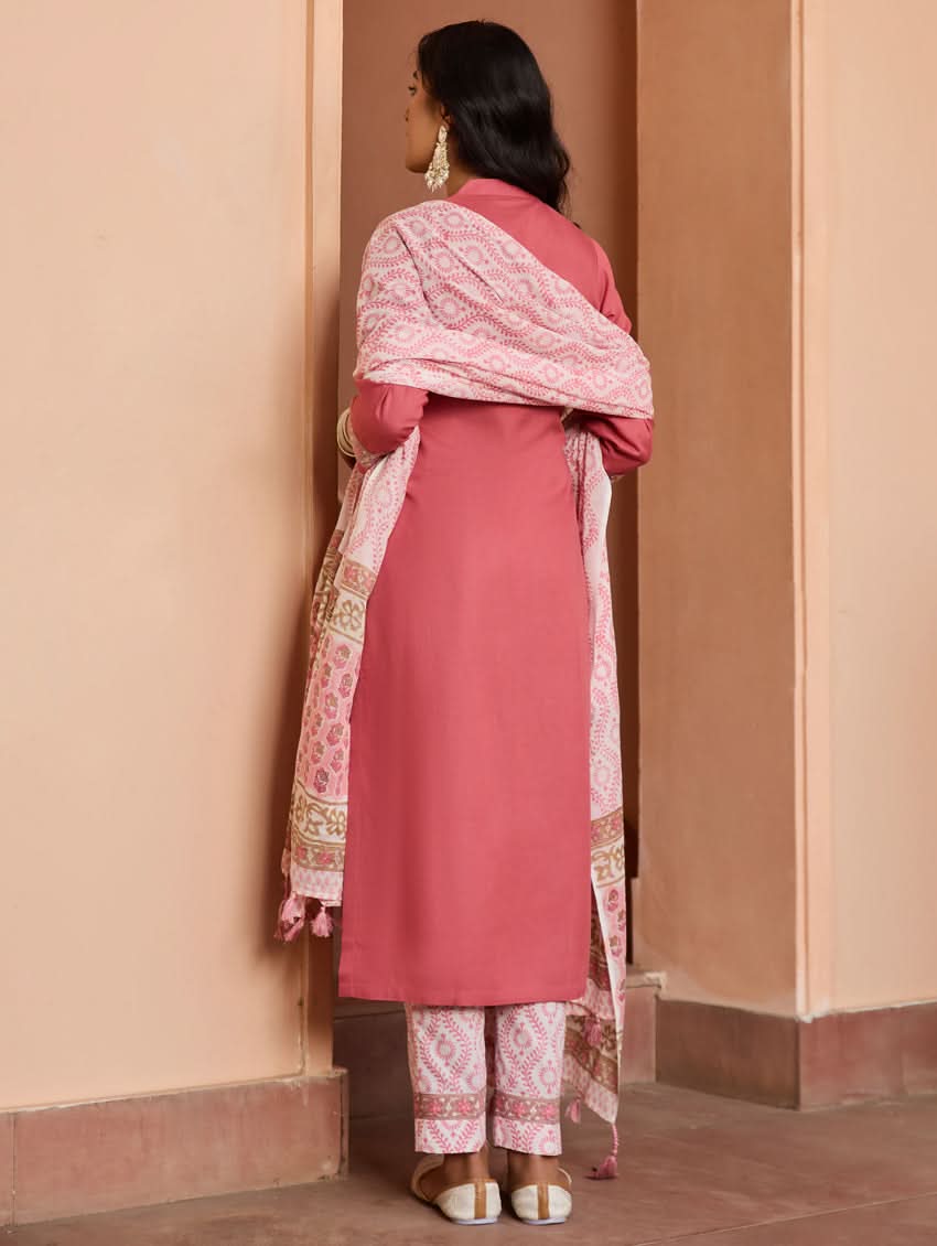 A woman wearing a pink embroidered rayon suit set with a kurta, pants, and a dupatta, standing in a doorway.