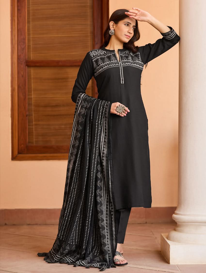 A woman wearing a suit set with embroidered details, consisting of a black kurta with a ban collar, three-fourth sleeves, and side slits, and black pants with a partially elasticated waistband. She is also wearing a printed mul dupatta with the suit set.