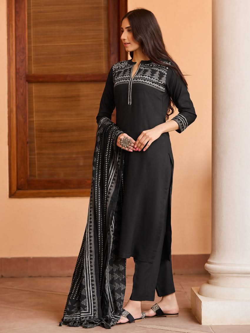 A woman wearing a suit set with embroidered details, consisting of a black kurta with a ban collar, three-fourth sleeves, and side slits, and black pants with a partially elasticated waistband. She is also wearing a printed mul dupatta with the suit set.