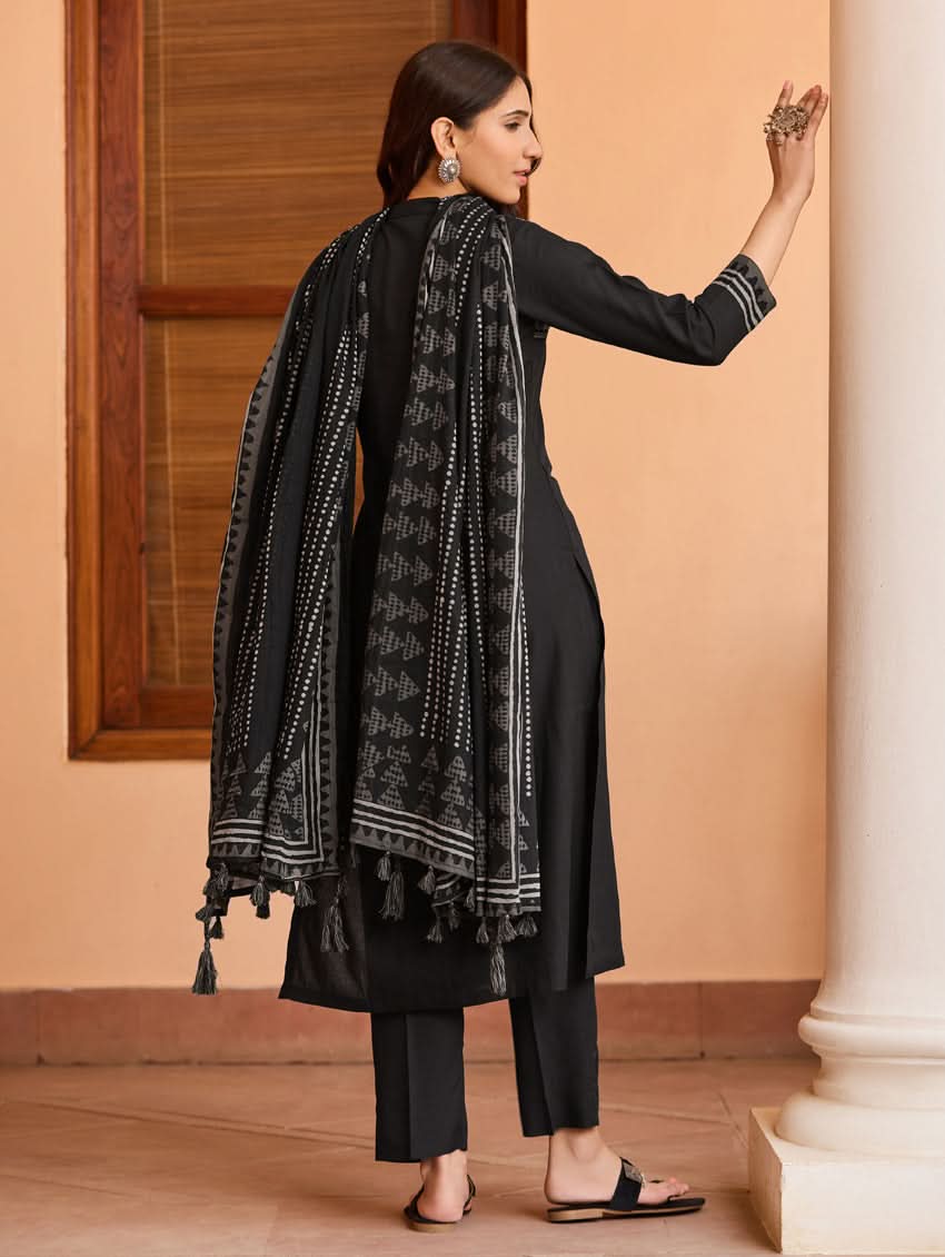 A woman wearing a suit set with embroidered details, consisting of a black kurta with a ban collar, three-fourth sleeves, and side slits, and black pants with a partially elasticated waistband. She is also wearing a printed mul dupatta with the suit set.