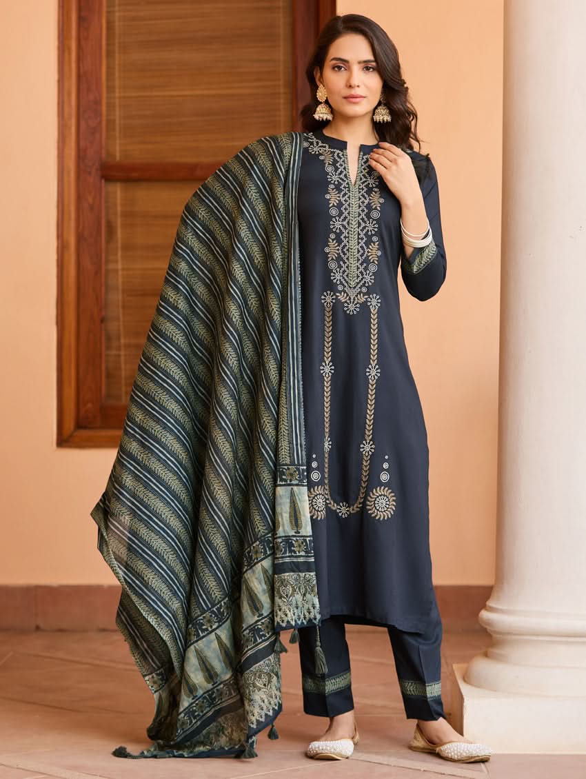 A woman wearing a traditional suit set with embroidered details, consisting of a black kurta with a ban collar, three-fourth sleeves, and side slits, and black pants with a partially elasticated waistband. She is also wearing a printed mul dupatta with the suit set.