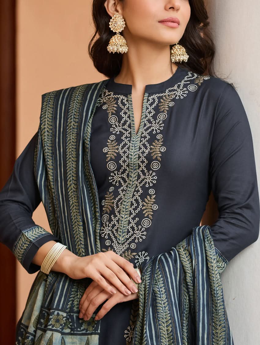 A woman wearing a traditional suit set with embroidered details, consisting of a black kurta with a ban collar, three-fourth sleeves, and side slits, and black pants with a partially elasticated waistband. She is also wearing a printed mul dupatta with the suit set.