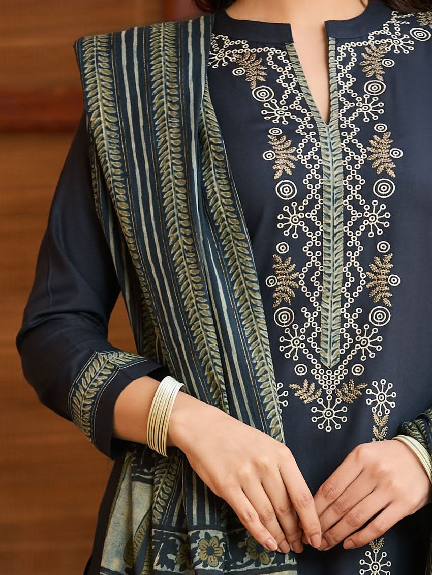 A woman wearing a traditional suit set with embroidered details, consisting of a black kurta with a ban collar, three-fourth sleeves, and side slits, and black pants with a partially elasticated waistband. She is also wearing a printed mul dupatta with the suit set.