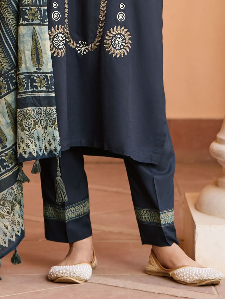 A woman wearing a traditional suit set with embroidered details, consisting of a black kurta with a ban collar, three-fourth sleeves, and side slits, and black pants with a partially elasticated waistband. She is also wearing a printed mul dupatta with the suit set.