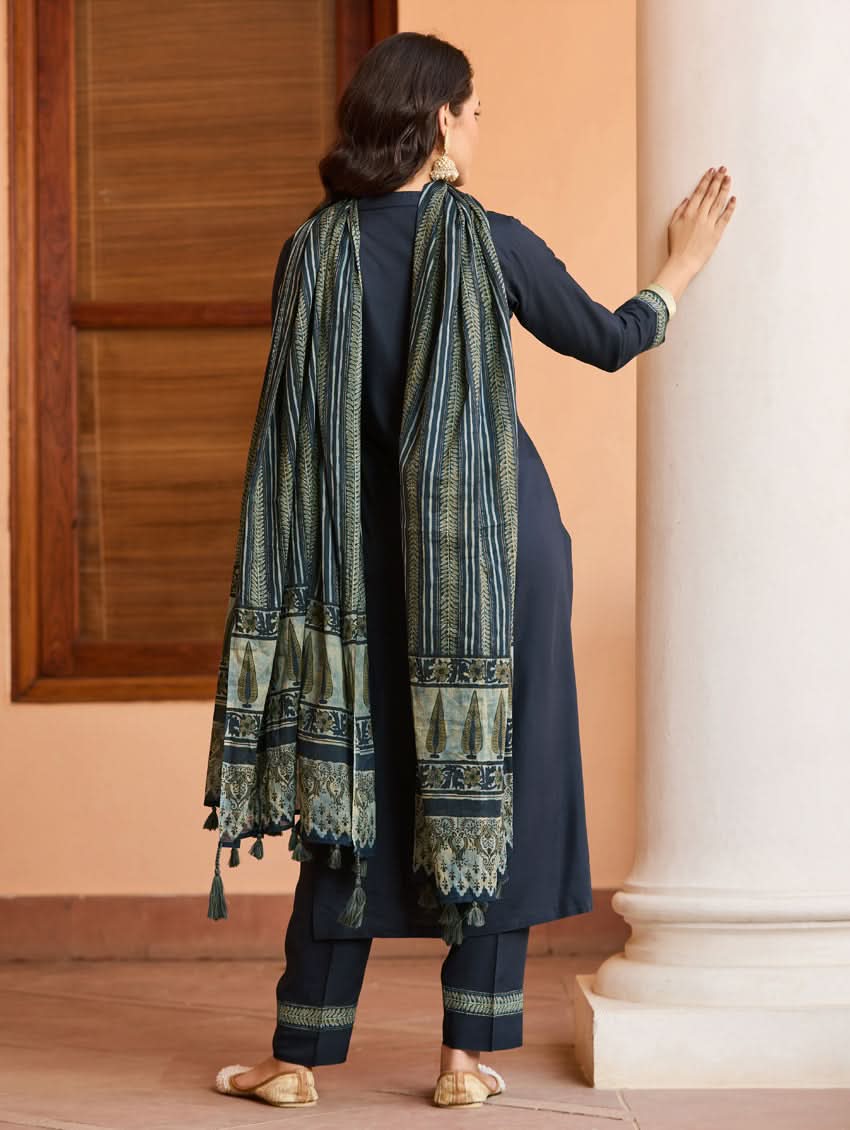A woman wearing a traditional suit set with embroidered details, consisting of a black kurta with a ban collar, three-fourth sleeves, and side slits, and black pants with a partially elasticated waistband. She is also wearing a printed mul dupatta with the suit set.