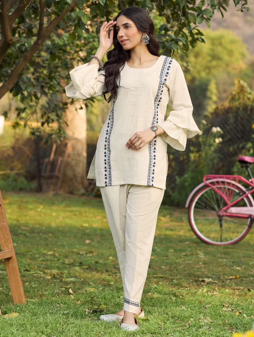 A woman standing outdoors wearing a beige embroidered viscose flex co-ord set with short kurta and pants, next to a pink bicycle.