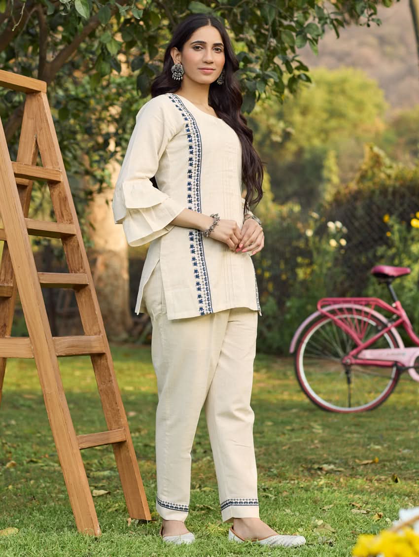 A woman standing outdoors wearing a beige embroidered viscose flex co-ord set with short kurta and pants, next to a pink bicycle.
