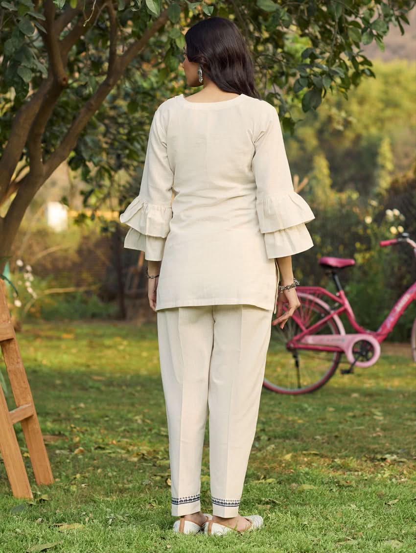 A woman standing outdoors wearing a beige embroidered viscose flex co-ord set with short kurta and pants, next to a pink bicycle.