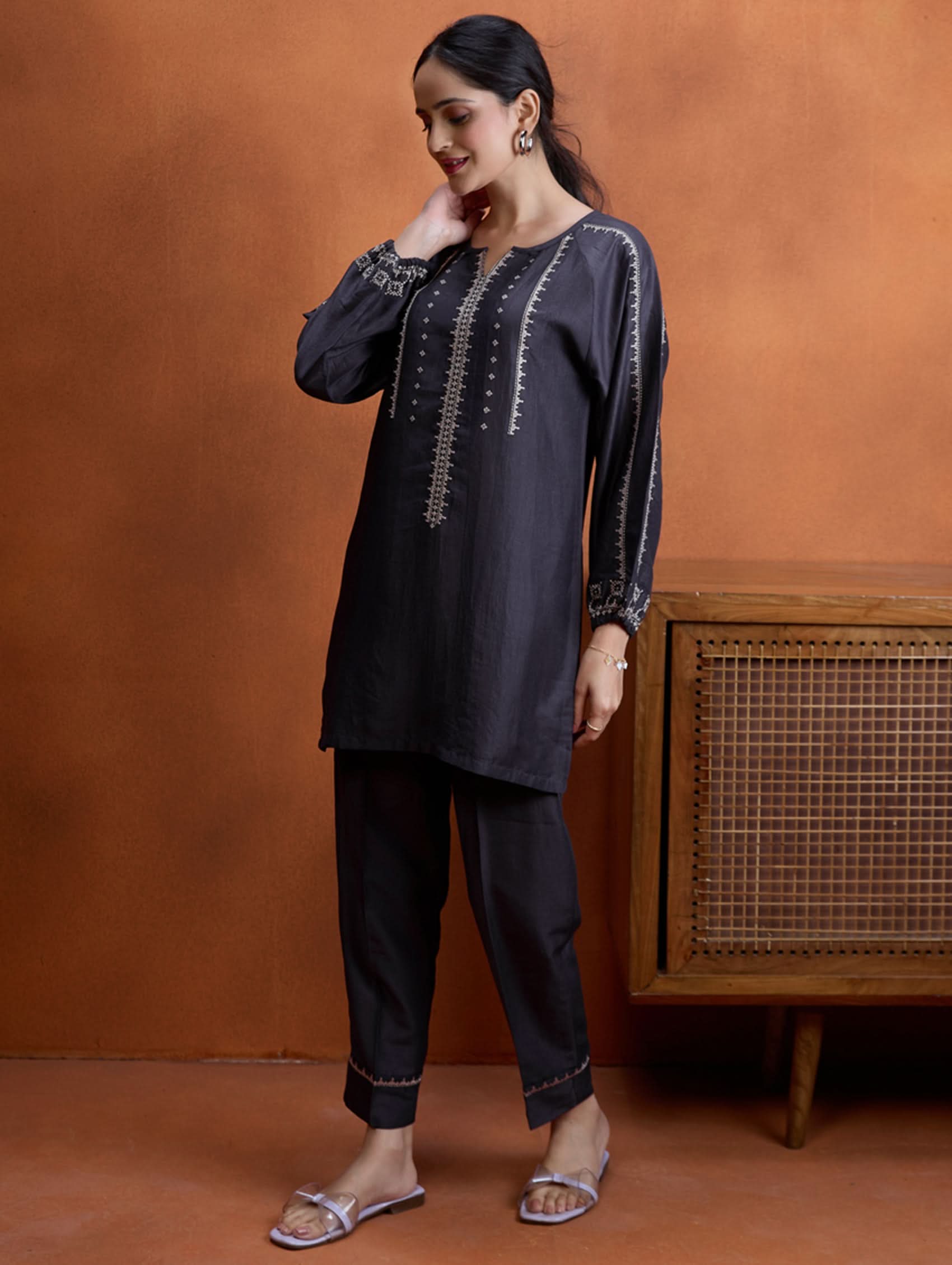 A model wearing a short kurta with a round collar and three-fourth sleeves, paired with pants that have partially elasticated waistband and pockets on either side. The fabric silk blend with computer embroidered