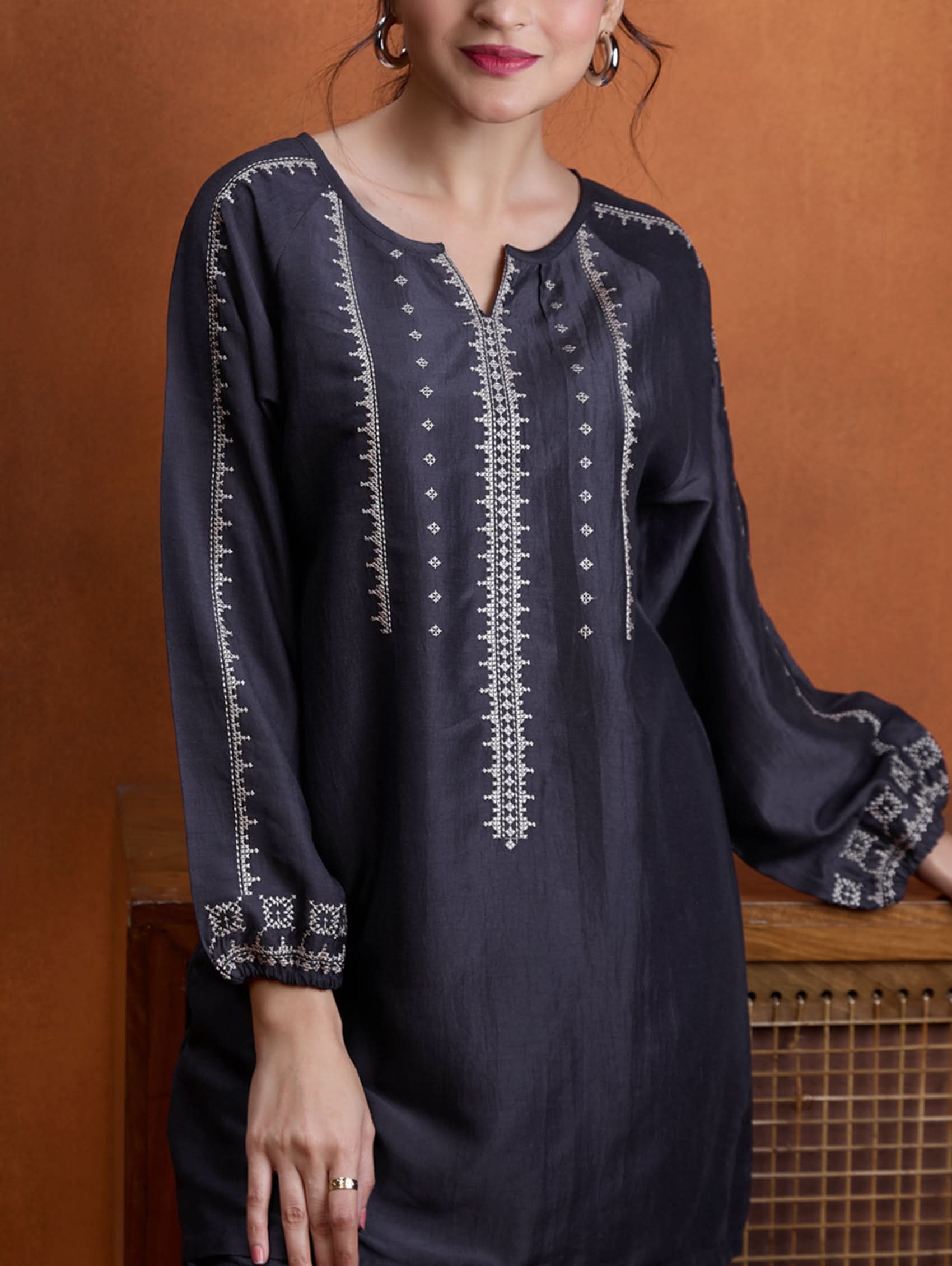 A model wearing a short kurta with a round collar and three-fourth sleeves, paired with pants that have partially elasticated waistband and pockets on either side. The fabric silk blend with computer embroidered