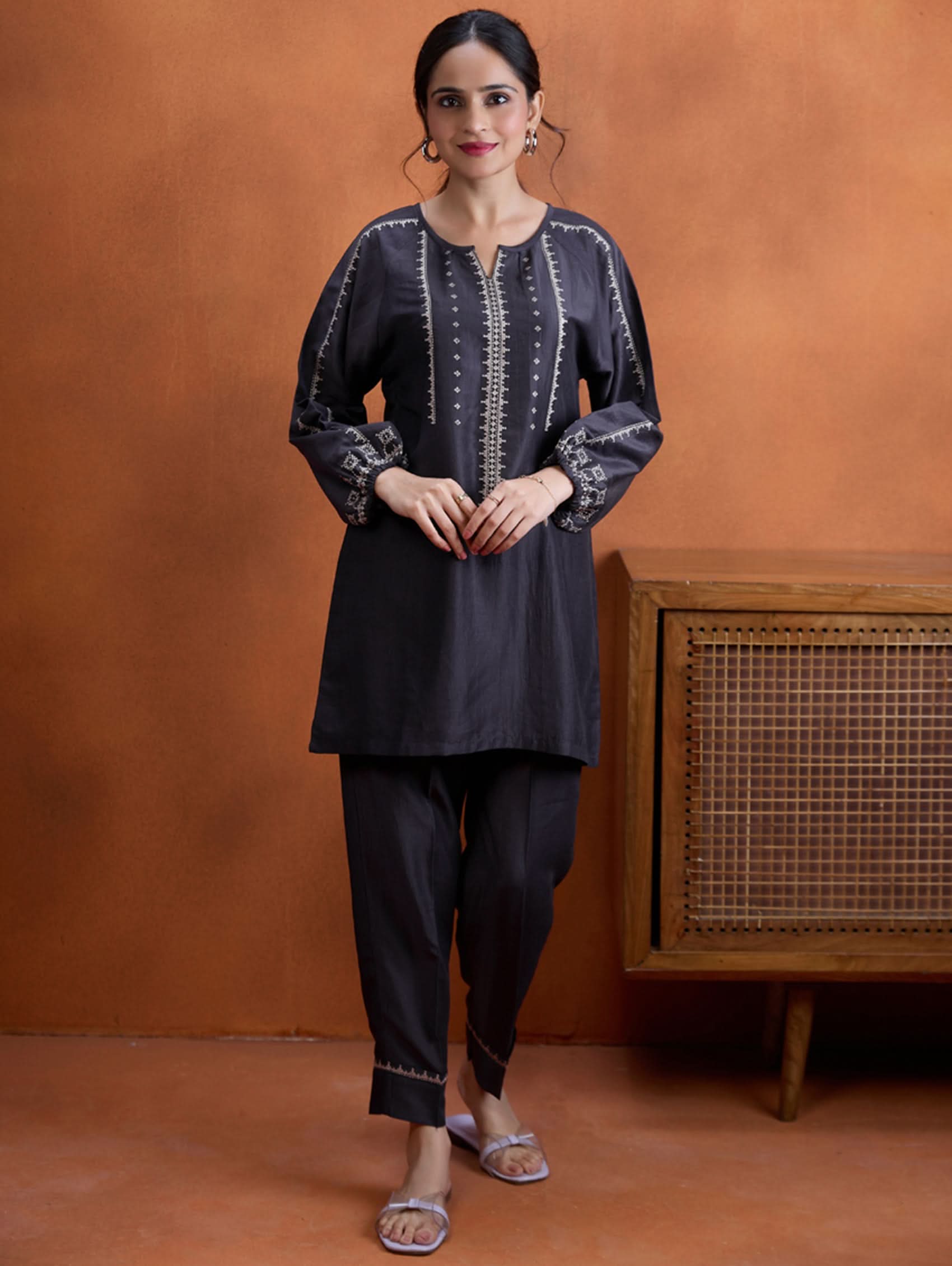 A model wearing a short kurta with a round collar and three-fourth sleeves, paired with pants that have partially elasticated waistband and pockets on either side. The fabric silk blend with computer embroidered