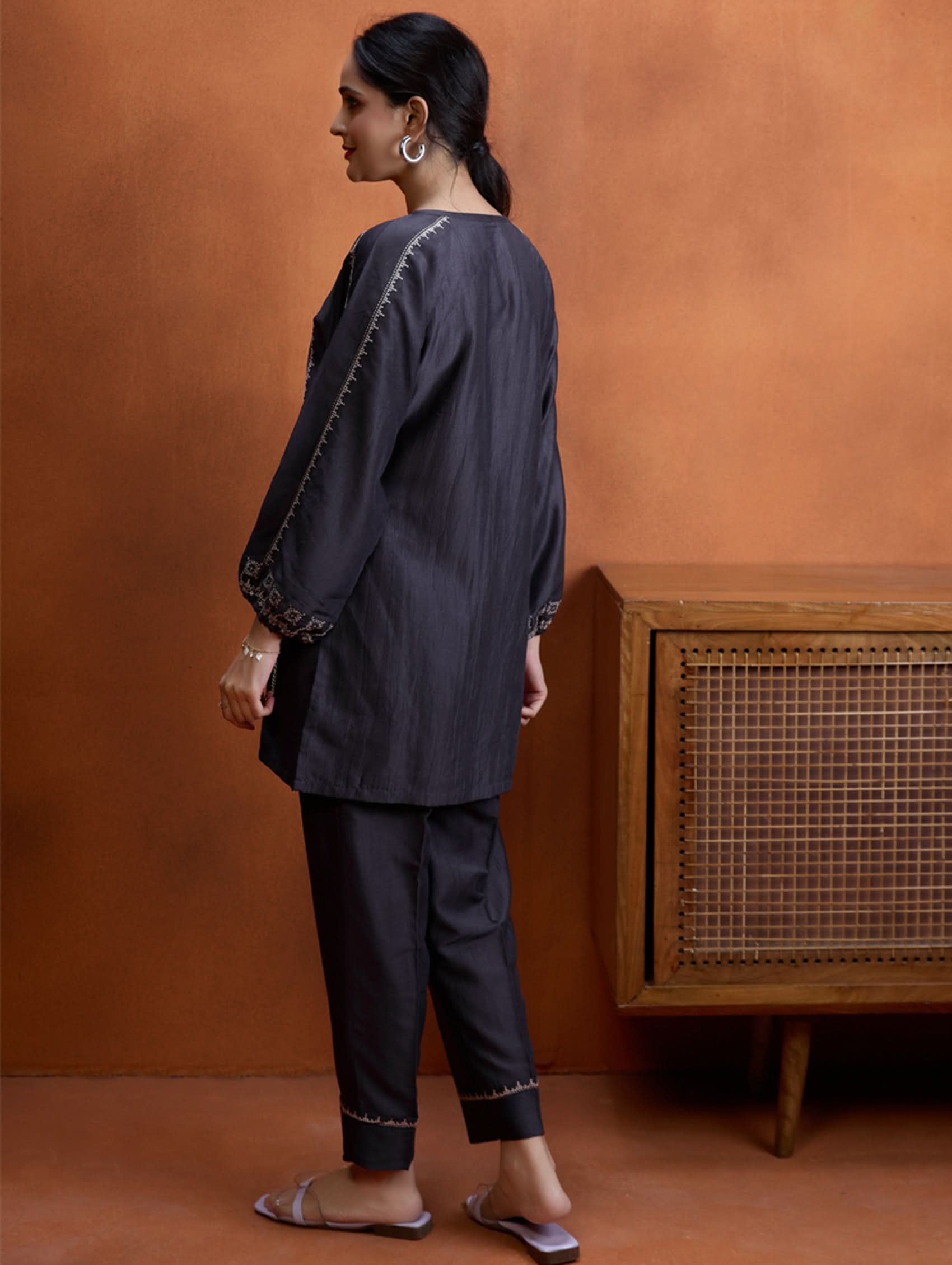 A model wearing a short kurta with a round collar and three-fourth sleeves, paired with pants that have partially elasticated waistband and pockets on either side. The fabric silk blend with computer embroidered