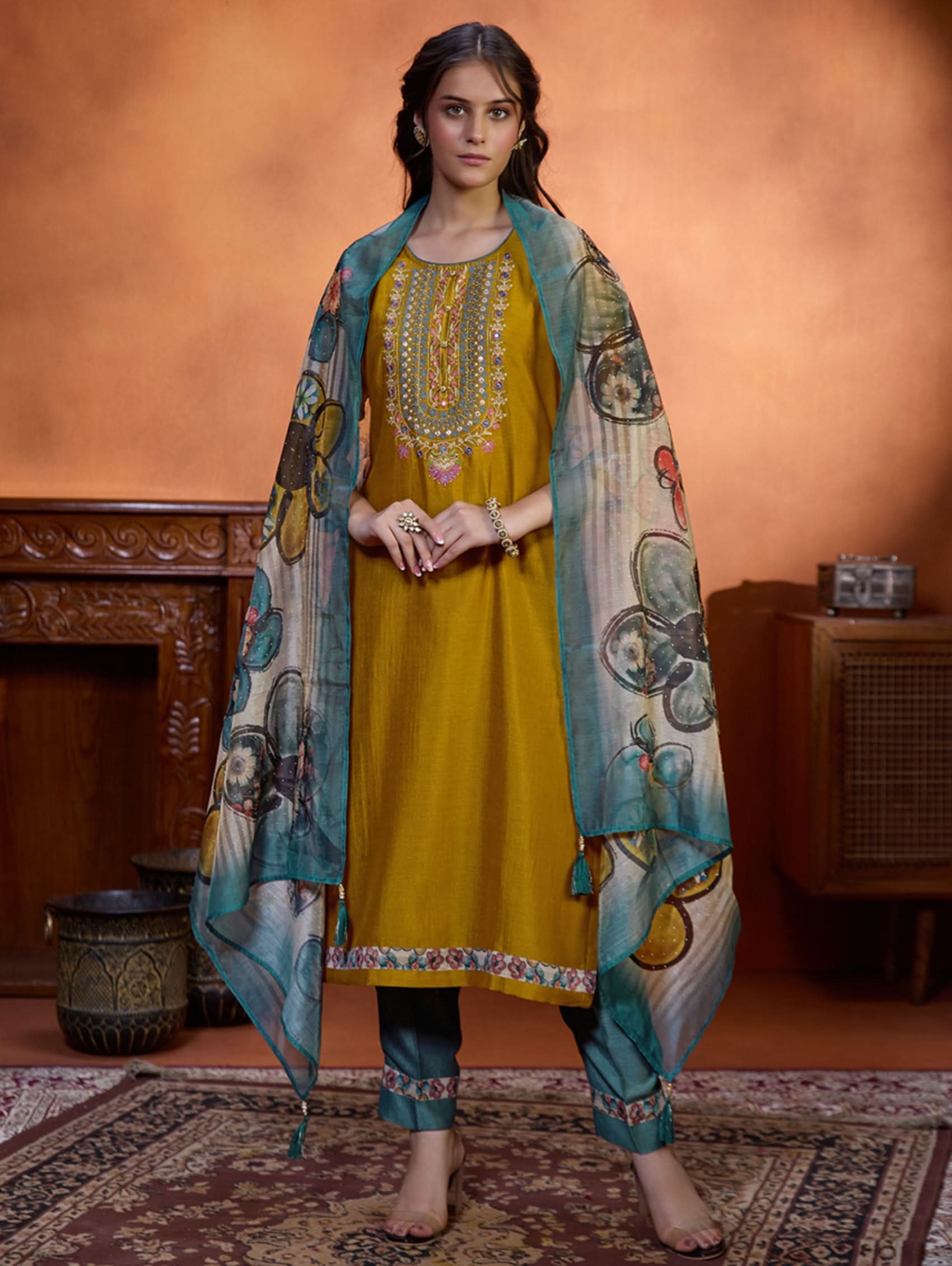 A woman wearing a traditional yellow kurta set with embroidered details and a dupatta with a digital printed chanderi silk design, standing in a room with an orange backdrop.