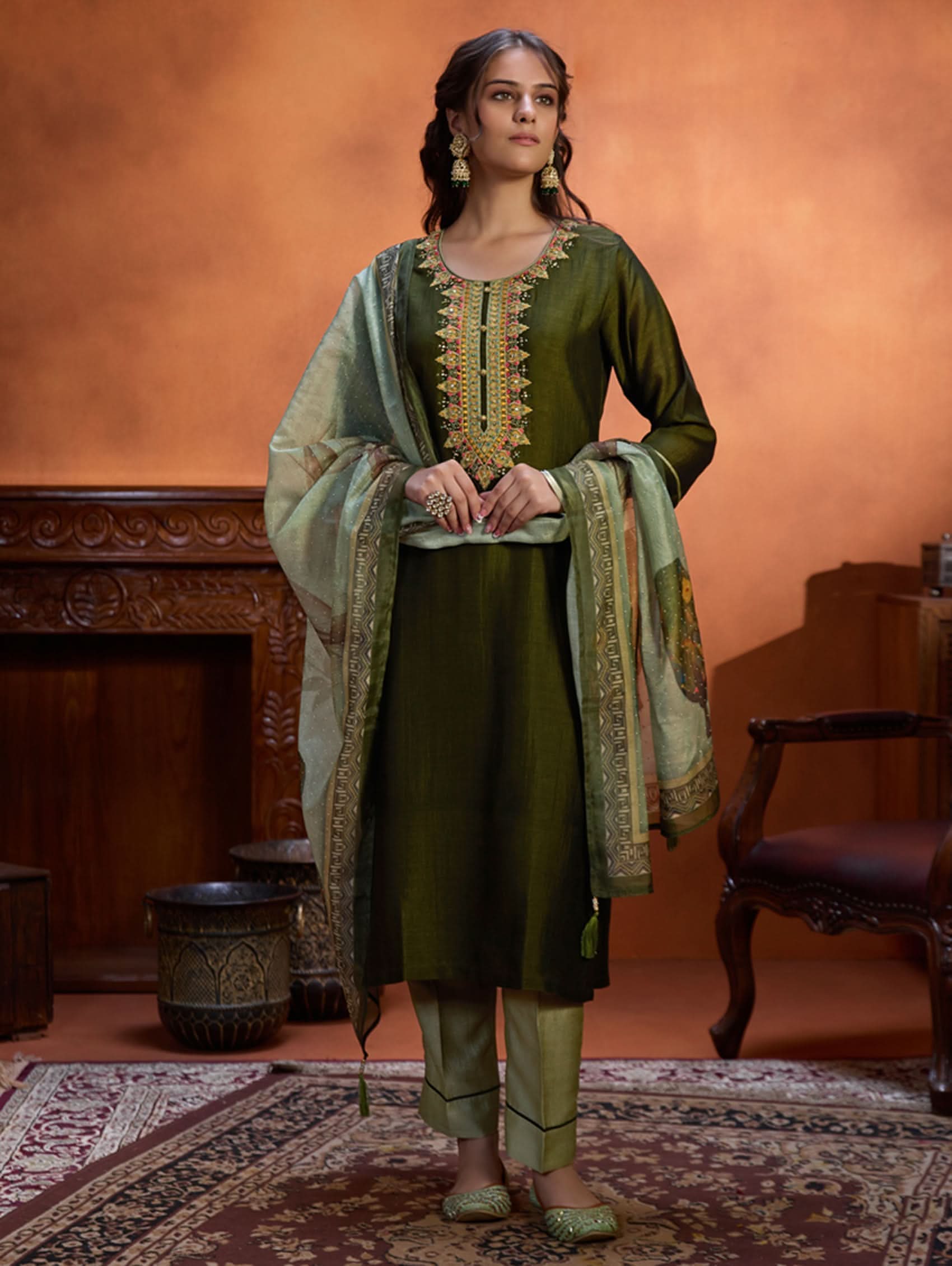 A woman wearing an embroidered viscose silk suit set with a green kurta and pants, a dupatta, and green earrings, standing in a room with an orange backdrop.