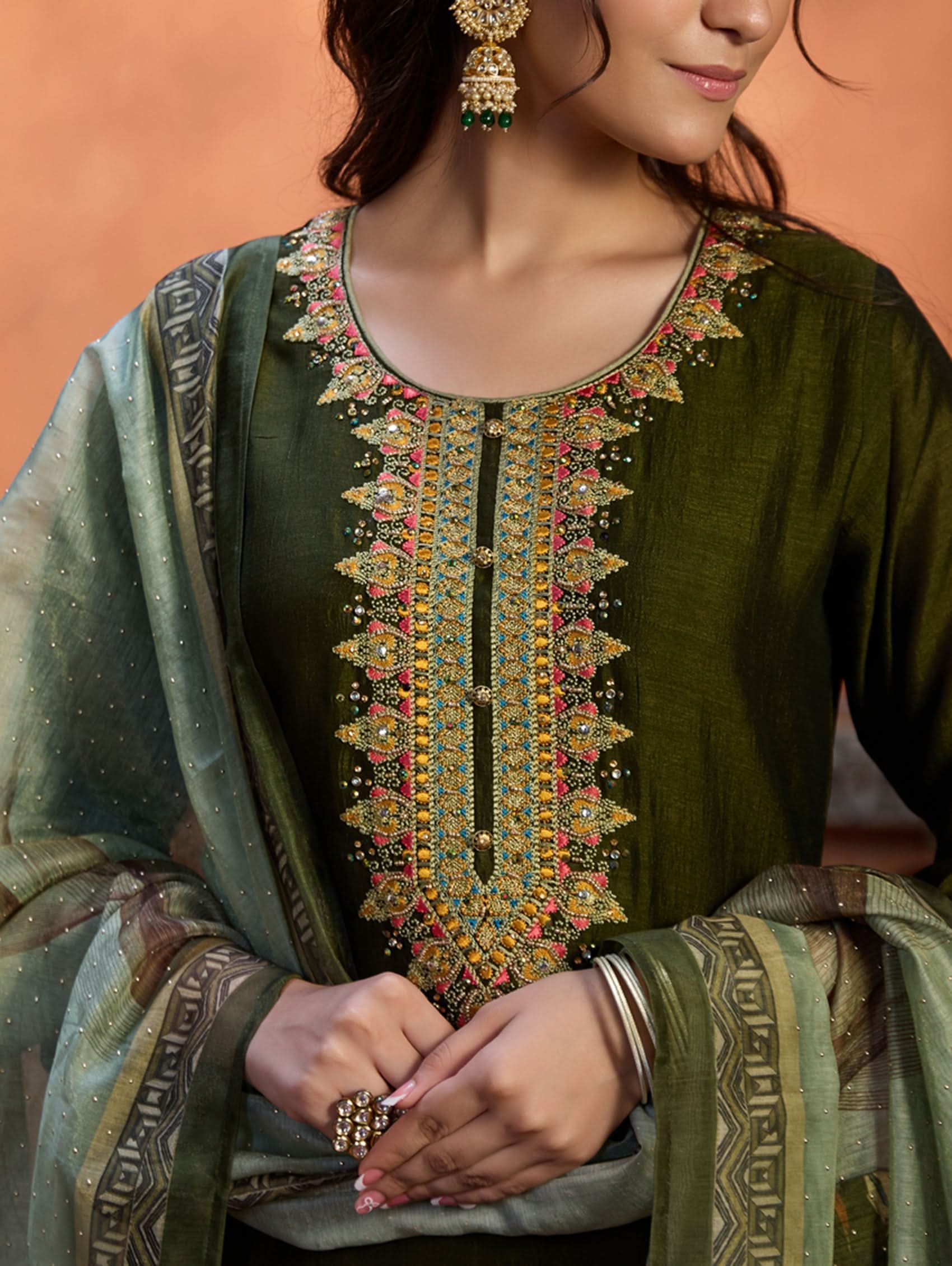 A woman wearing an embroidered viscose silk suit set with a green kurta and pants, a dupatta, and green earrings, standing in a room with an orange backdrop.
