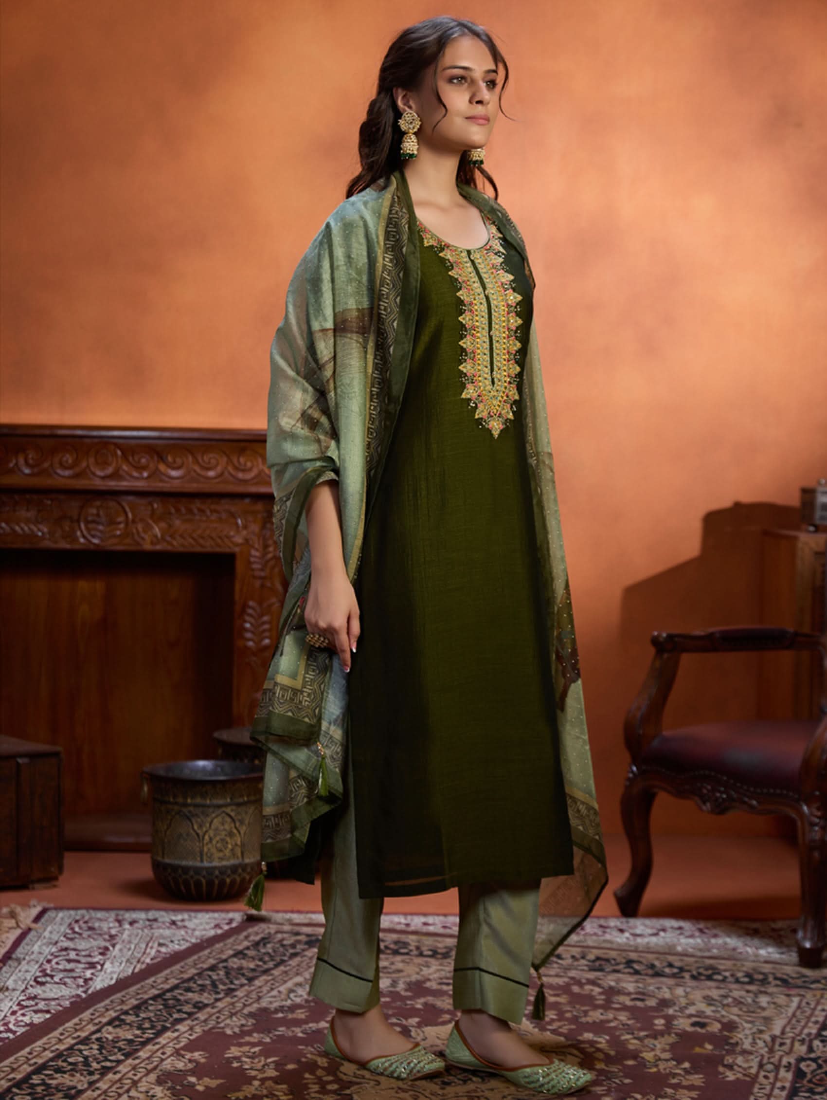 A woman wearing an embroidered viscose silk suit set with a green kurta and pants, a dupatta, and green earrings, standing in a room with an orange backdrop.