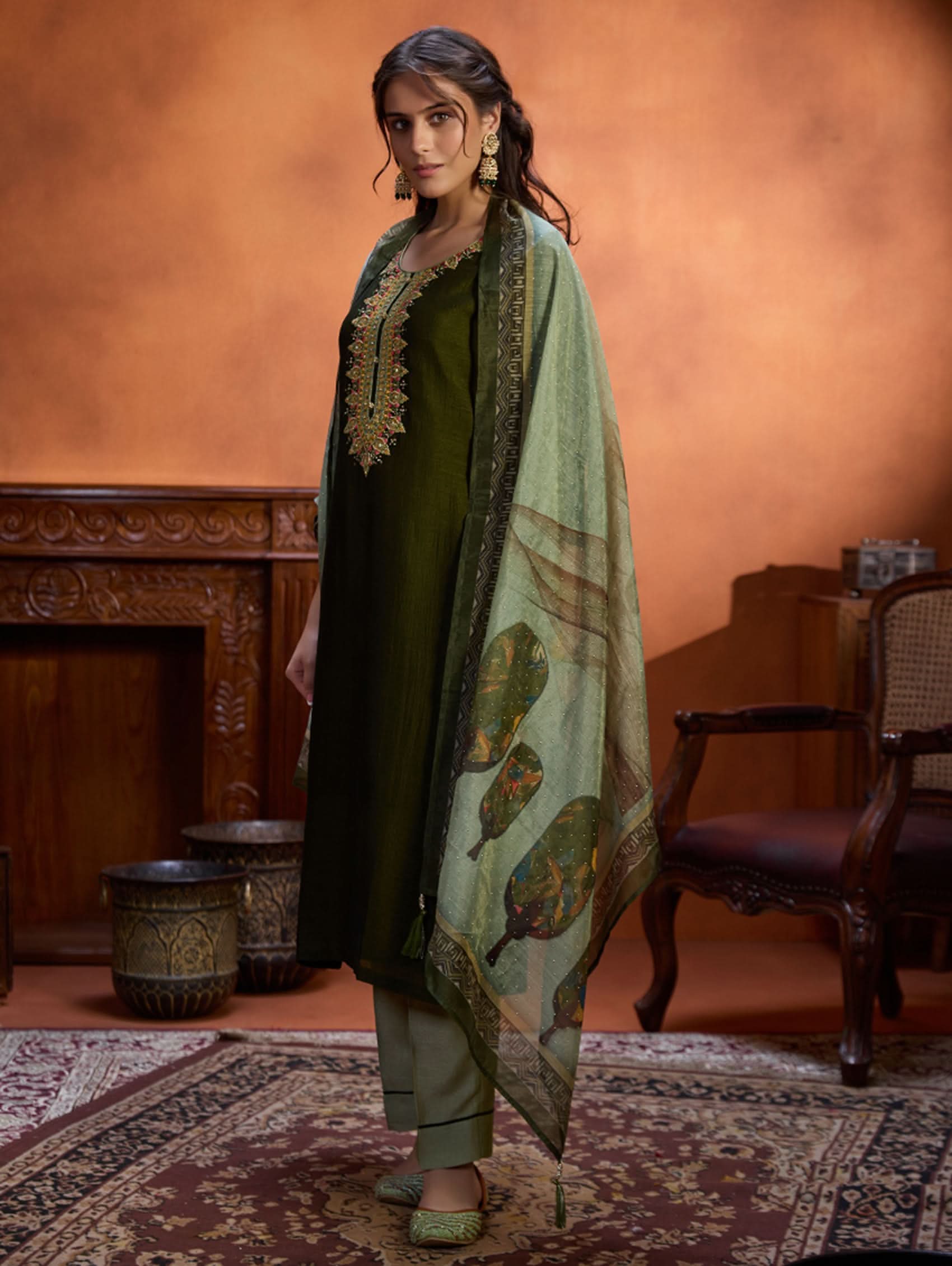 A woman wearing an embroidered viscose silk suit set with a green kurta and pants, a dupatta, and green earrings, standing in a room with an orange backdrop.