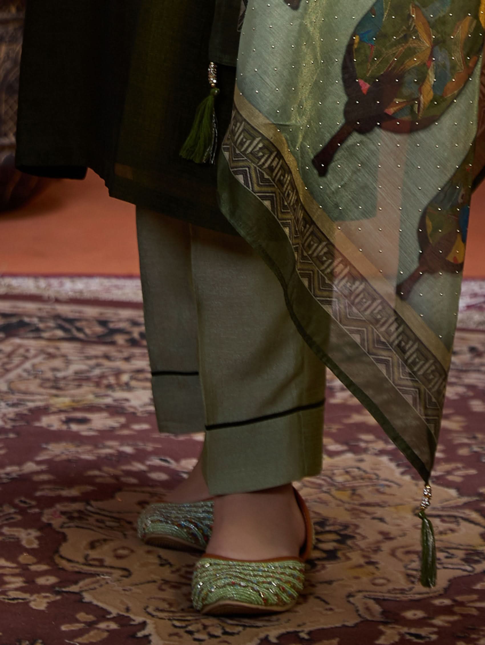 A woman wearing an embroidered viscose silk suit set with a green kurta and pants, a dupatta, and green earrings, standing in a room with an orange backdrop.