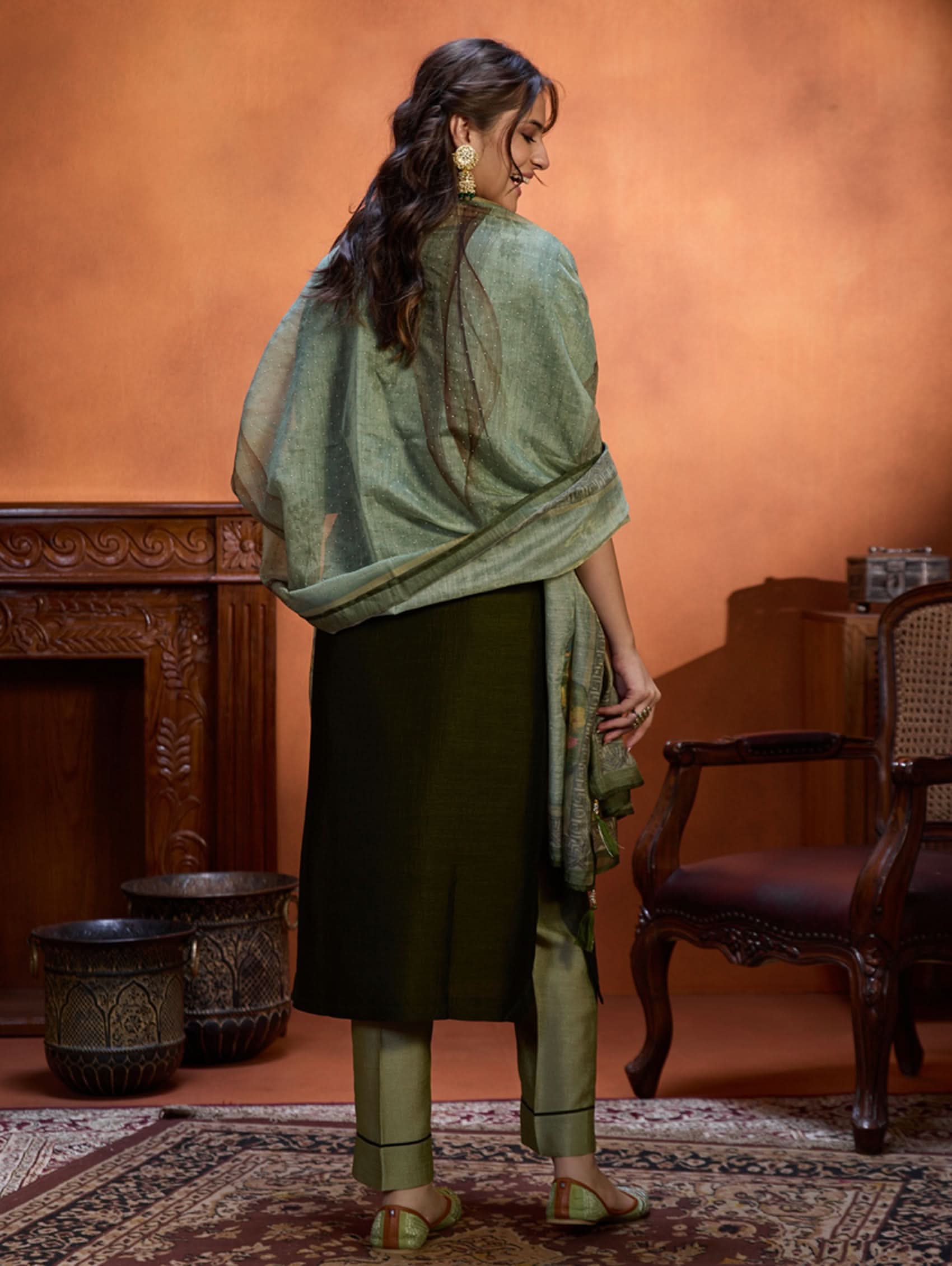 A woman wearing an embroidered viscose silk suit set with a green kurta and pants, a dupatta, and green earrings, standing in a room with an orange backdrop.