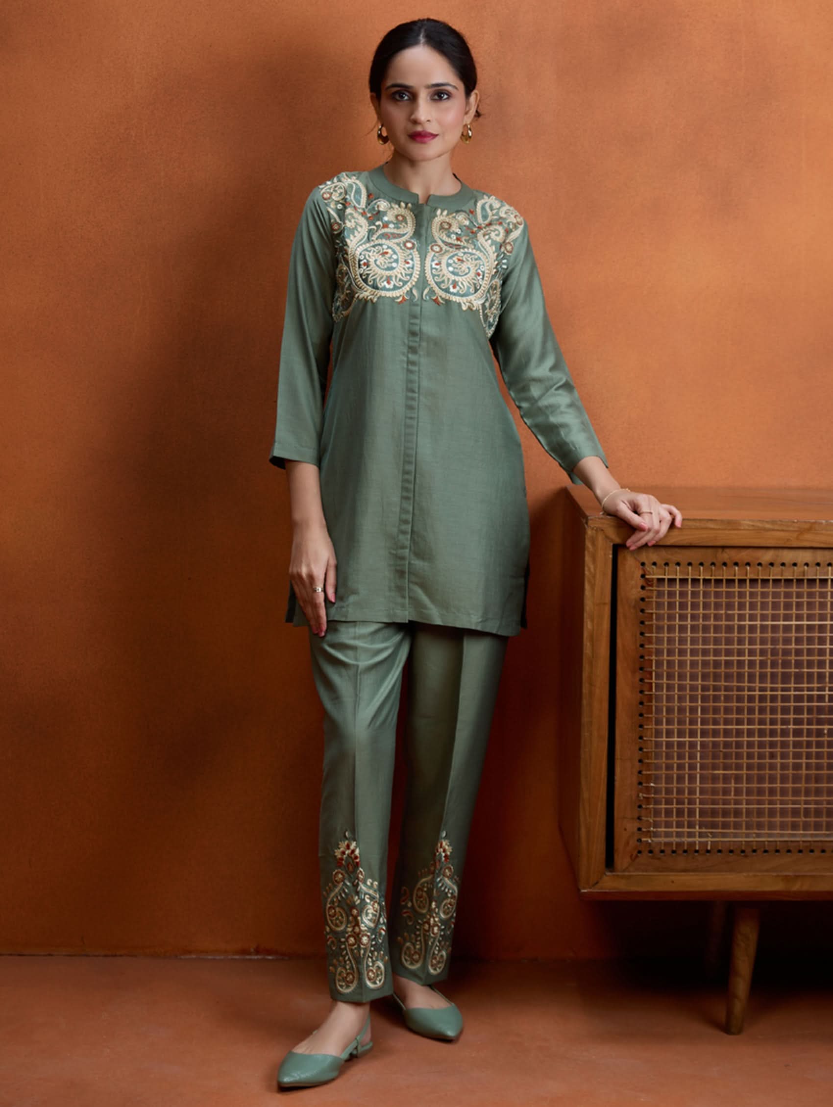 A woman standing next to a wooden column, wearing an embroidered green viscose silk co-ord set with a short kurta and pants, featuring a ban collar and three-quarter sleeves.