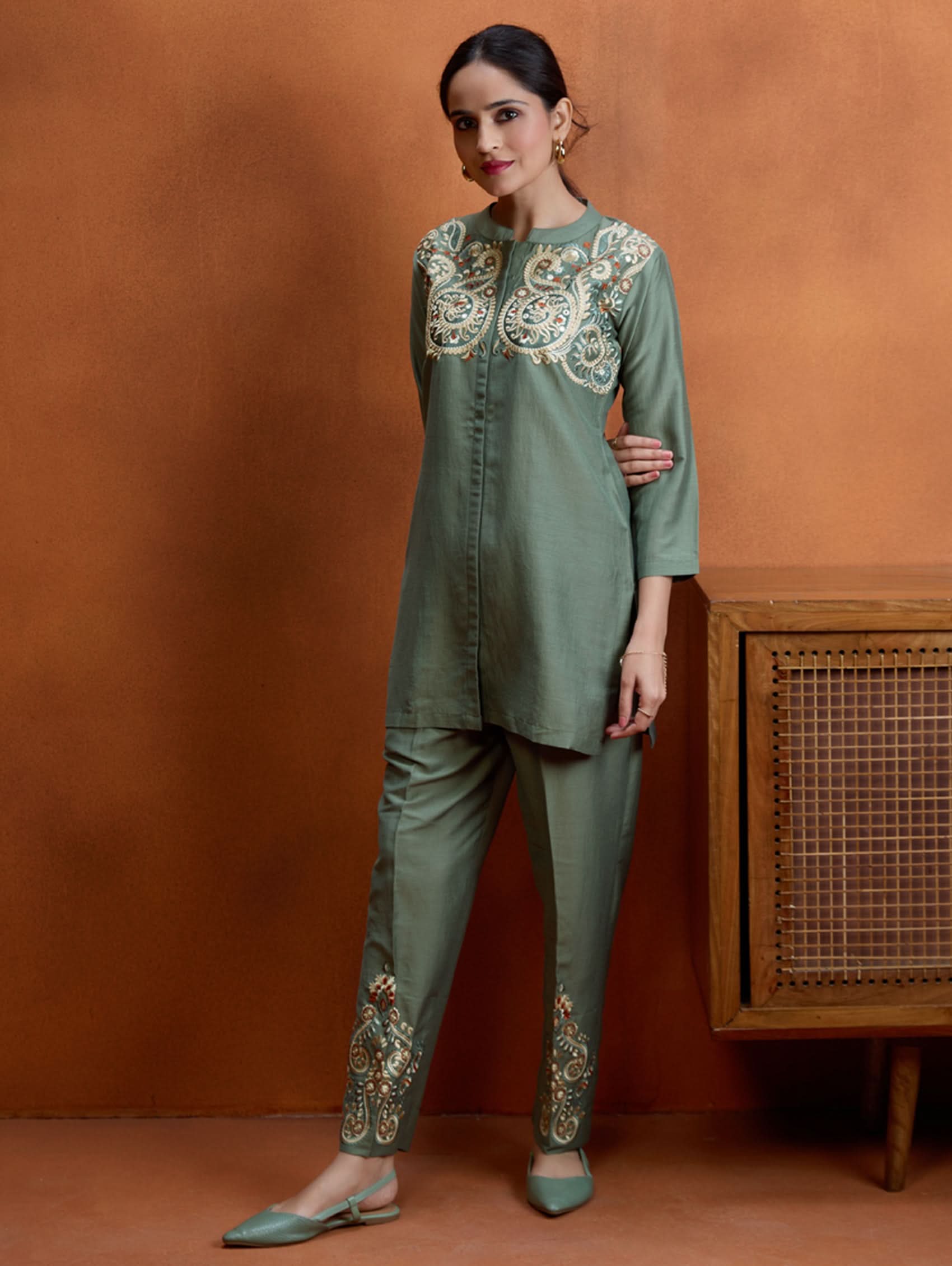 A woman standing next to a wooden column, wearing an embroidered green viscose silk co-ord set with a short kurta and pants, featuring a ban collar and three-quarter sleeves.