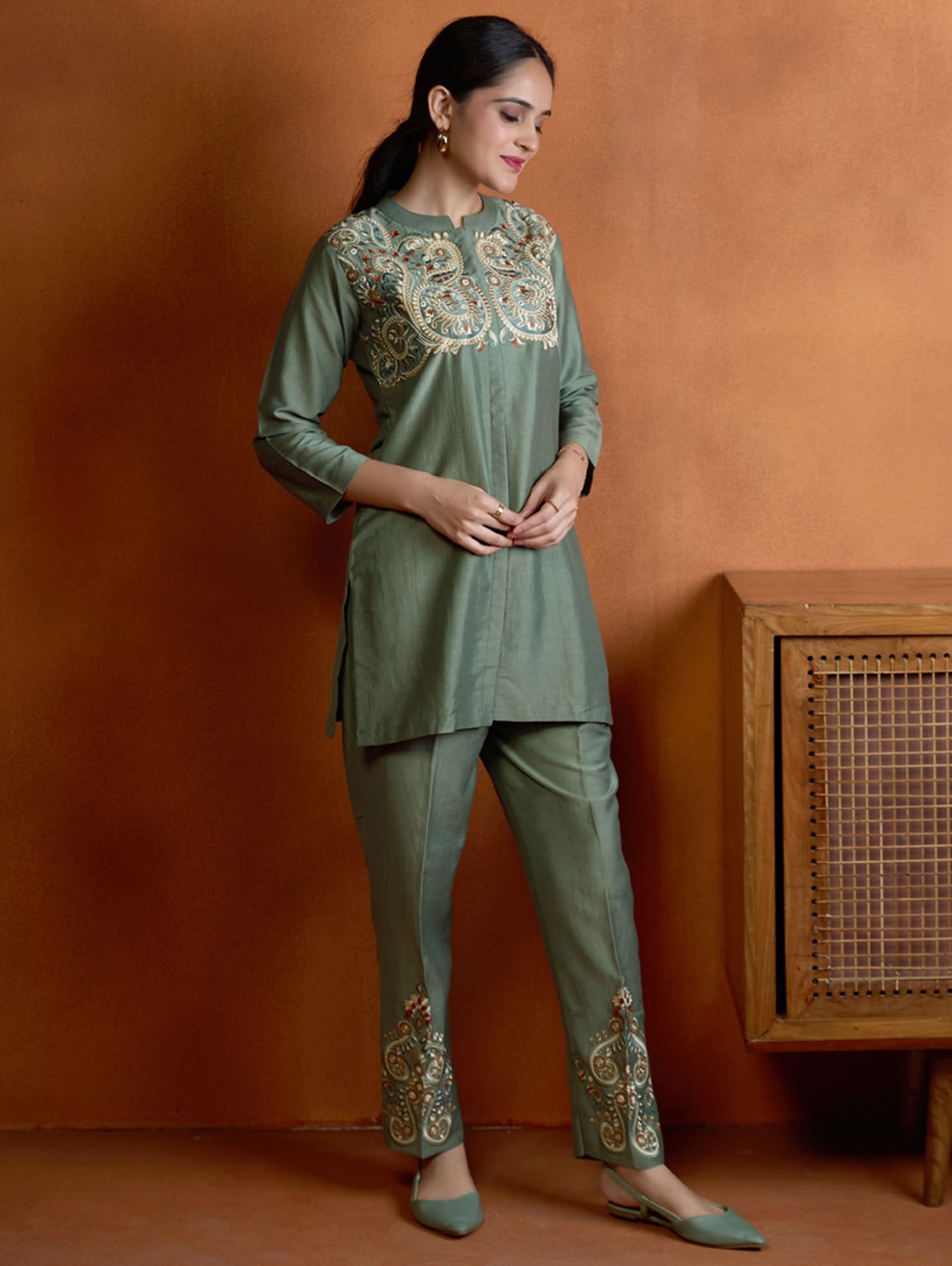 A woman standing next to a wooden column, wearing an embroidered green viscose silk co-ord set with a short kurta and pants, featuring a ban collar and three-quarter sleeves.