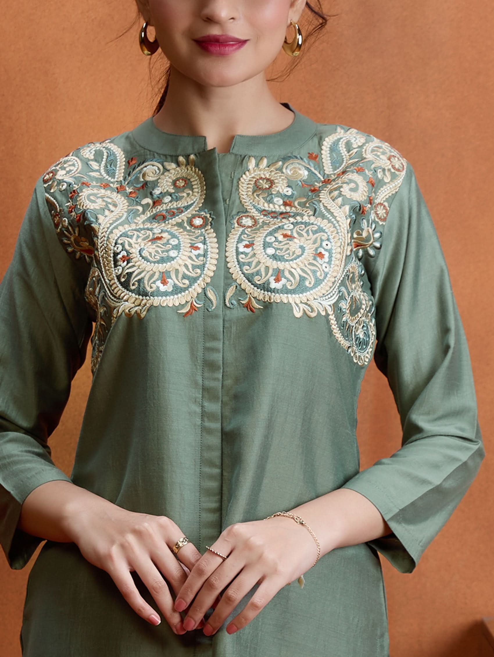 A woman standing next to a wooden column, wearing an embroidered green viscose silk co-ord set with a short kurta and pants, featuring a ban collar and three-quarter sleeves.