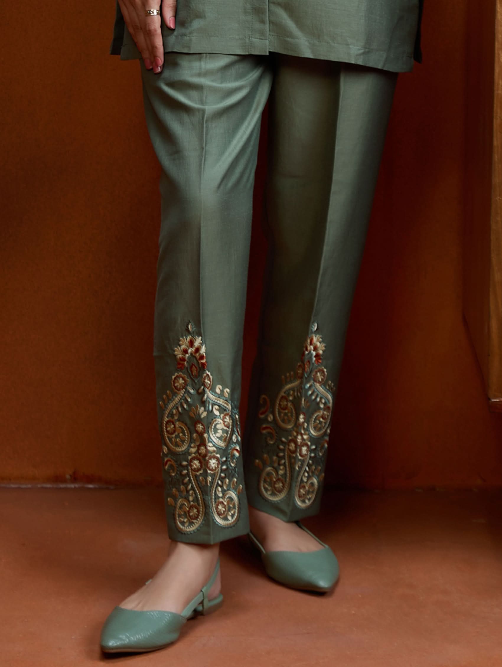 A woman standing next to a wooden column, wearing an embroidered green viscose silk co-ord set with a short kurta and pants, featuring a ban collar and three-quarter sleeves.