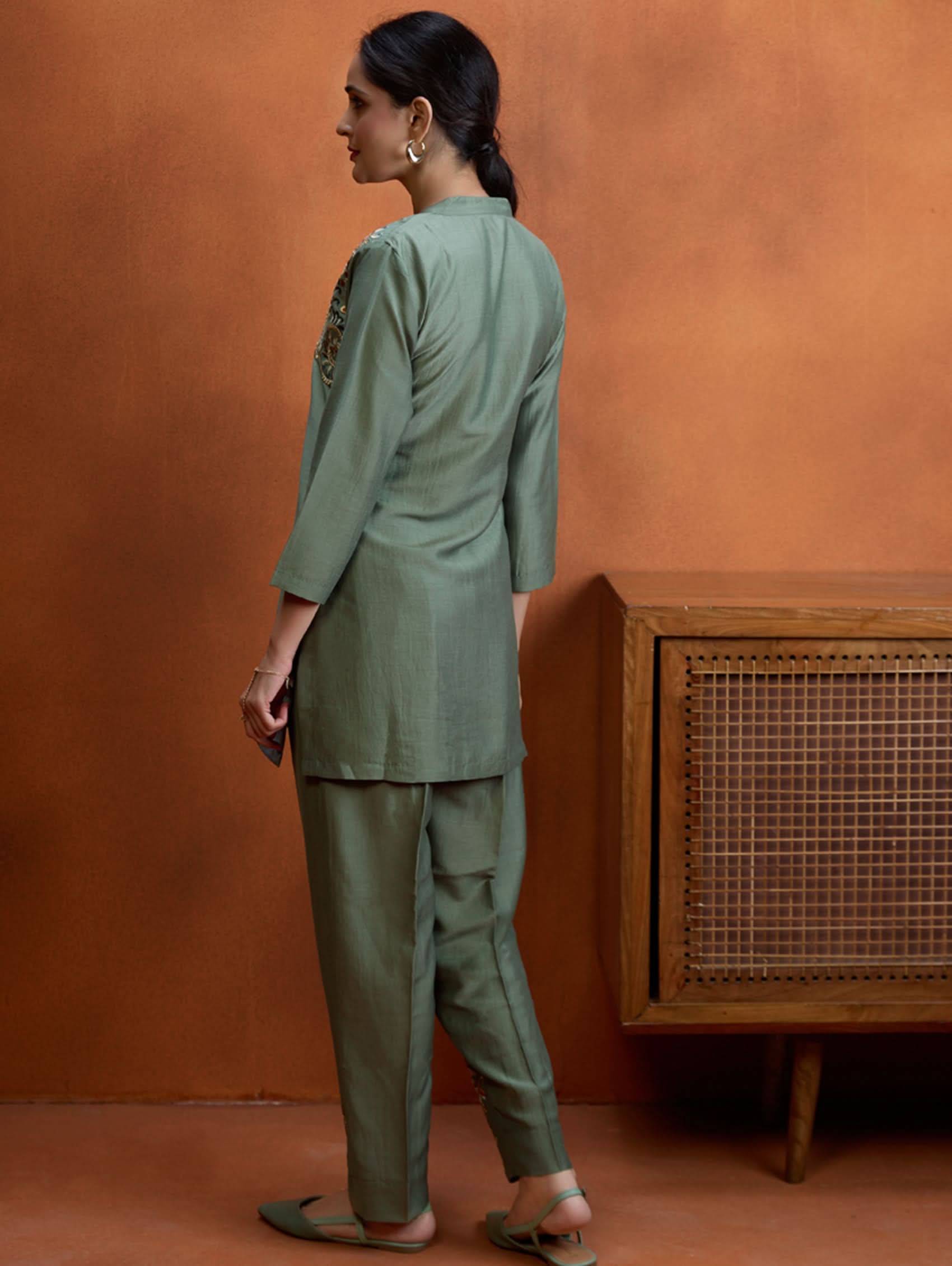 A woman standing next to a wooden column, wearing an embroidered green viscose silk co-ord set with a short kurta and pants, featuring a ban collar and three-quarter sleeves.