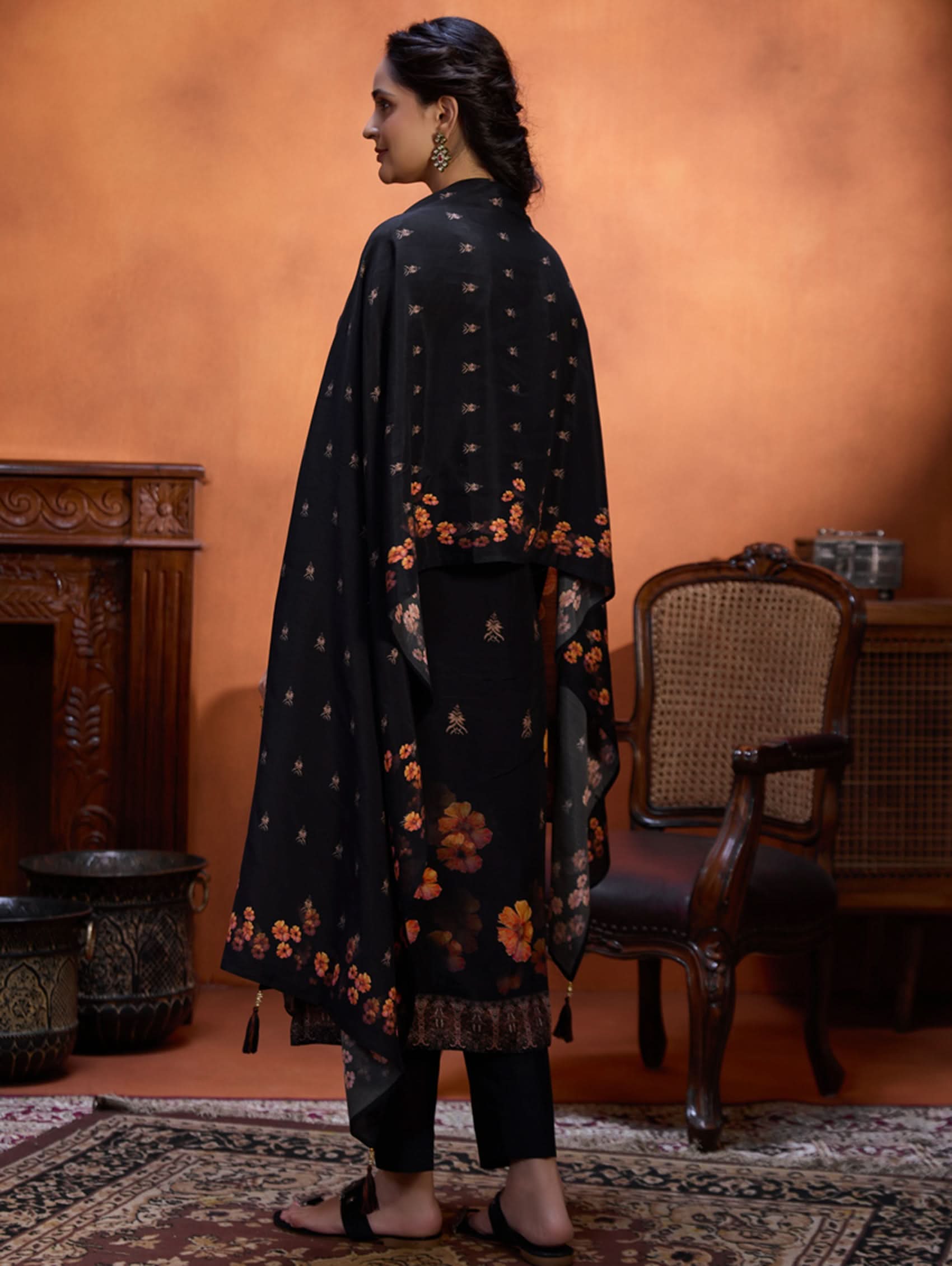 Woman wearing a black traditional outfit with floral patterns in a warm-toned room.