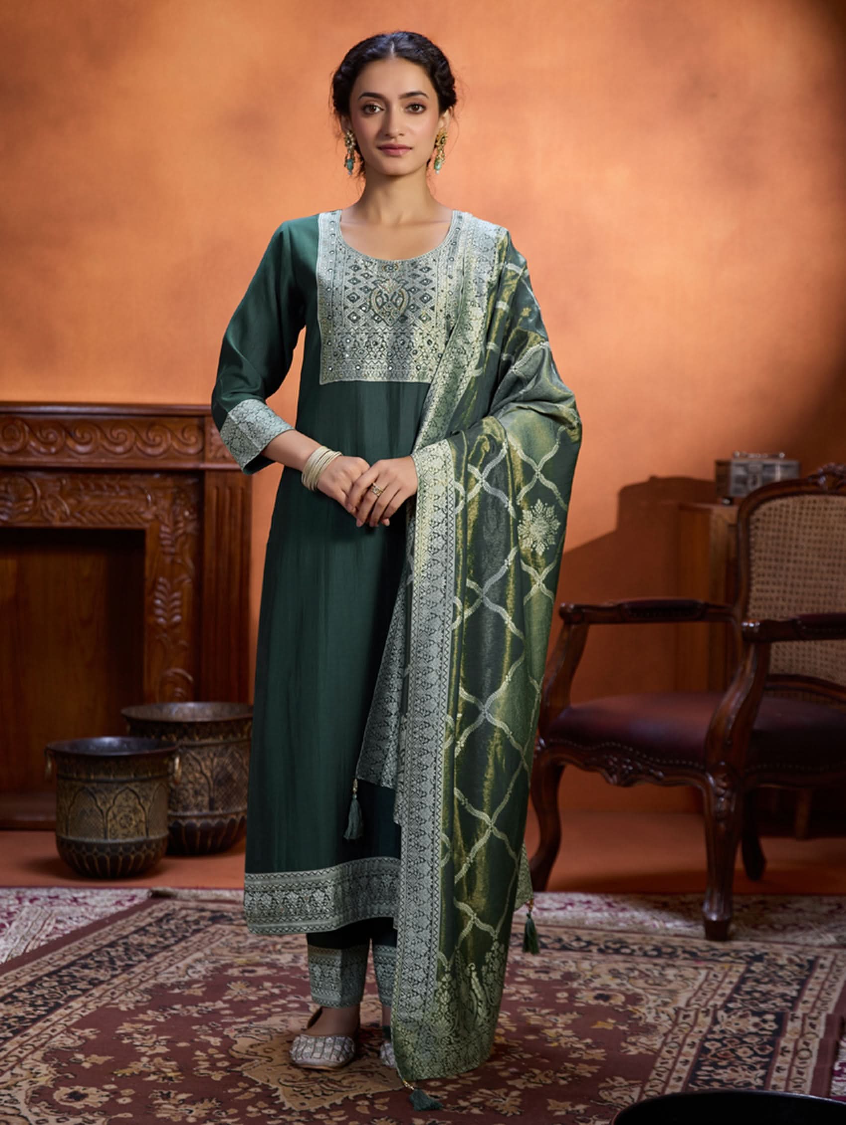 A woman wearing a green viscose silk suit set with a solid-coloured kurta, pants, and a jacquard weave shimmer silk dupatta, standing in a room with wooden furniture.