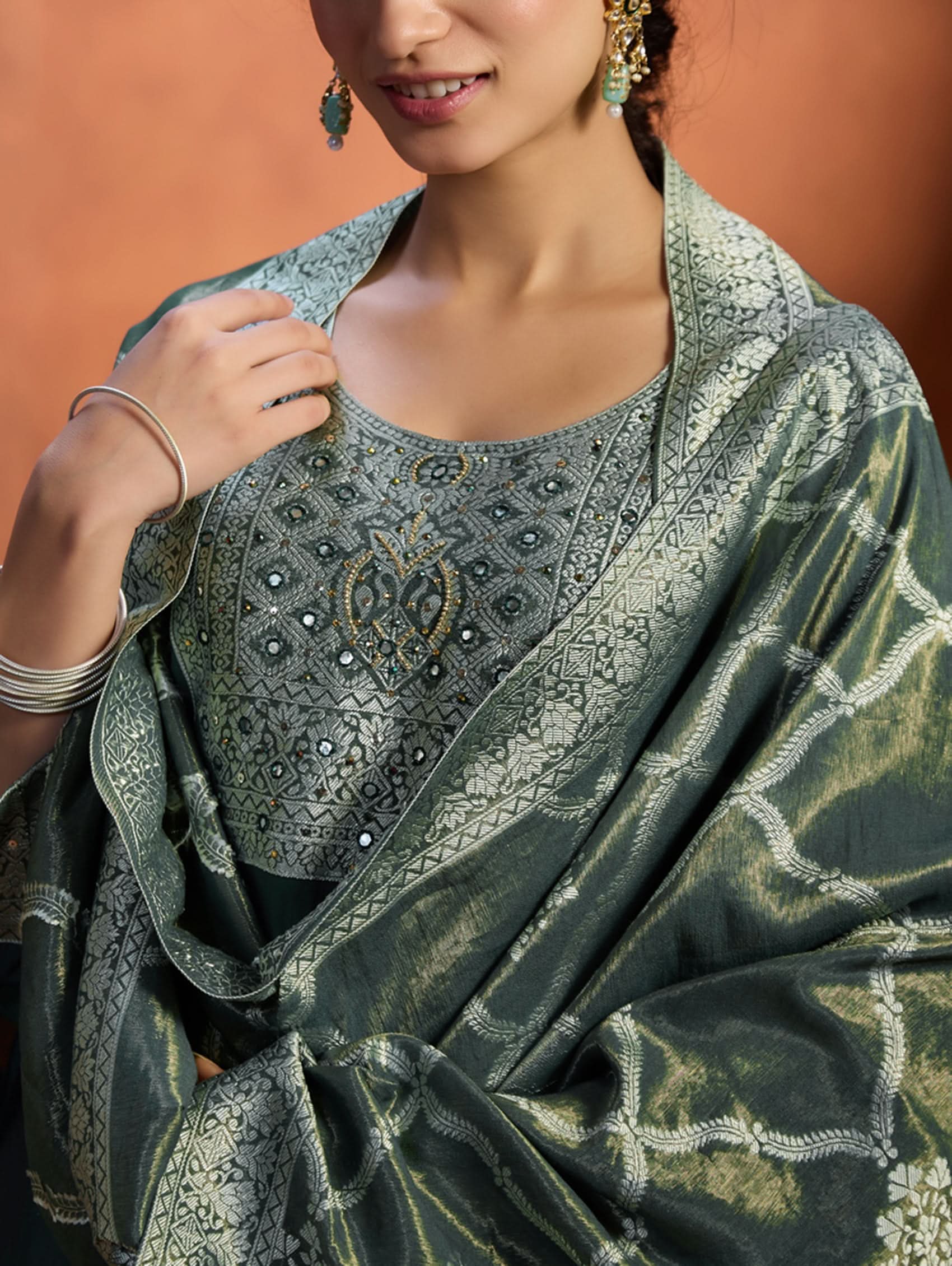 A woman wearing a green viscose silk suit set with a solid-coloured kurta, pants, and a jacquard weave shimmer silk dupatta, standing in a room with wooden furniture.