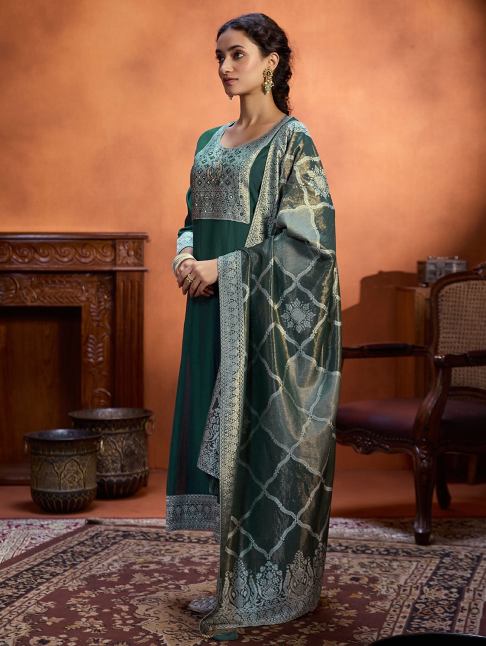 A woman wearing a green viscose silk suit set with a solid-coloured kurta, pants, and a jacquard weave shimmer silk dupatta, standing in a room with wooden furniture.