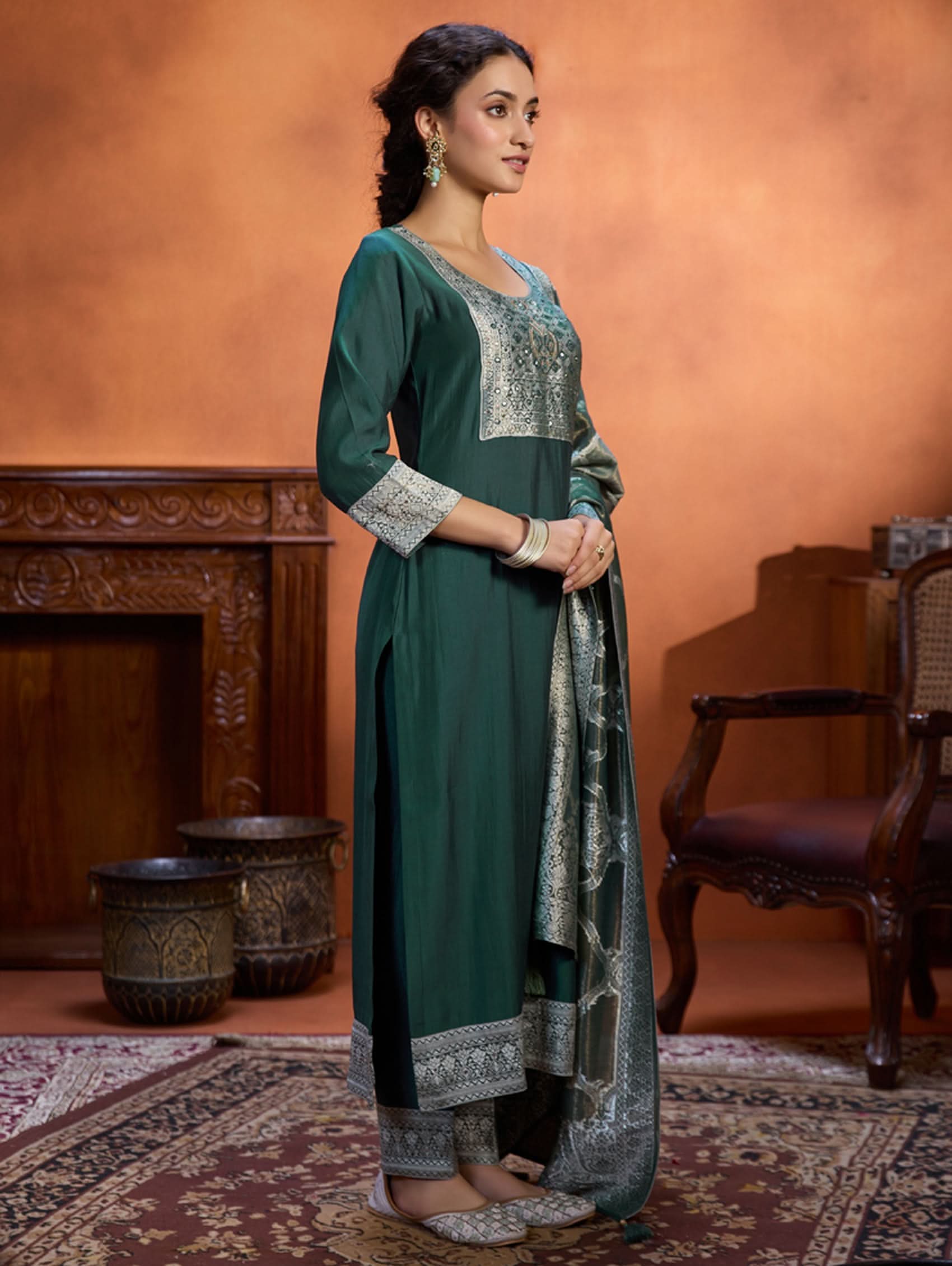 A woman wearing a green viscose silk suit set with a solid-coloured kurta, pants, and a jacquard weave shimmer silk dupatta, standing in a room with wooden furniture.