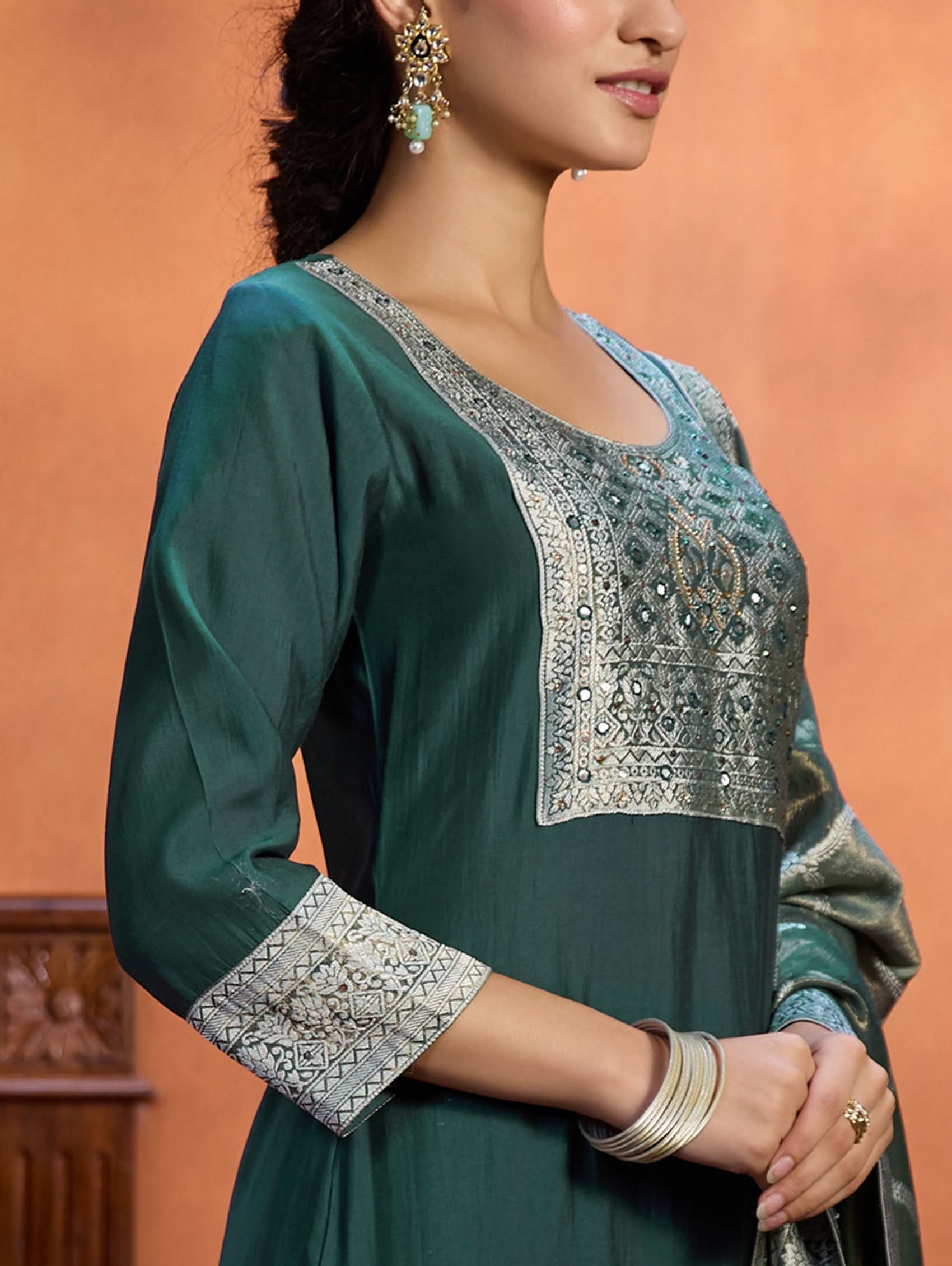 A woman wearing a green viscose silk suit set with a solid-coloured kurta, pants, and a jacquard weave shimmer silk dupatta, standing in a room with wooden furniture.