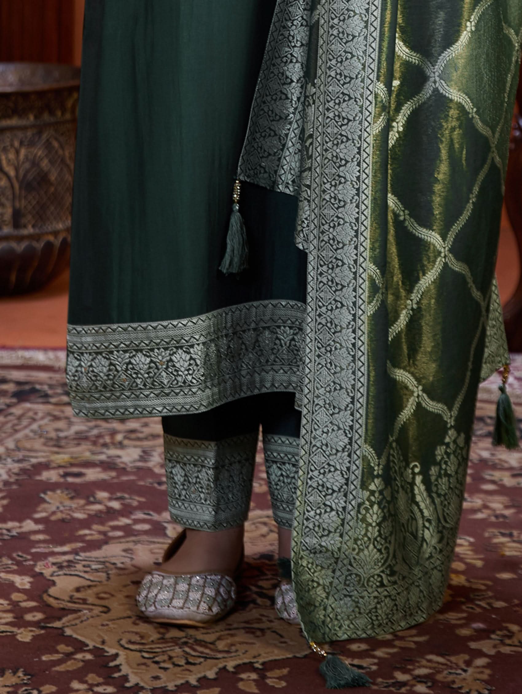 A woman wearing a green viscose silk suit set with a solid-coloured kurta, pants, and a jacquard weave shimmer silk dupatta, standing in a room with wooden furniture.
