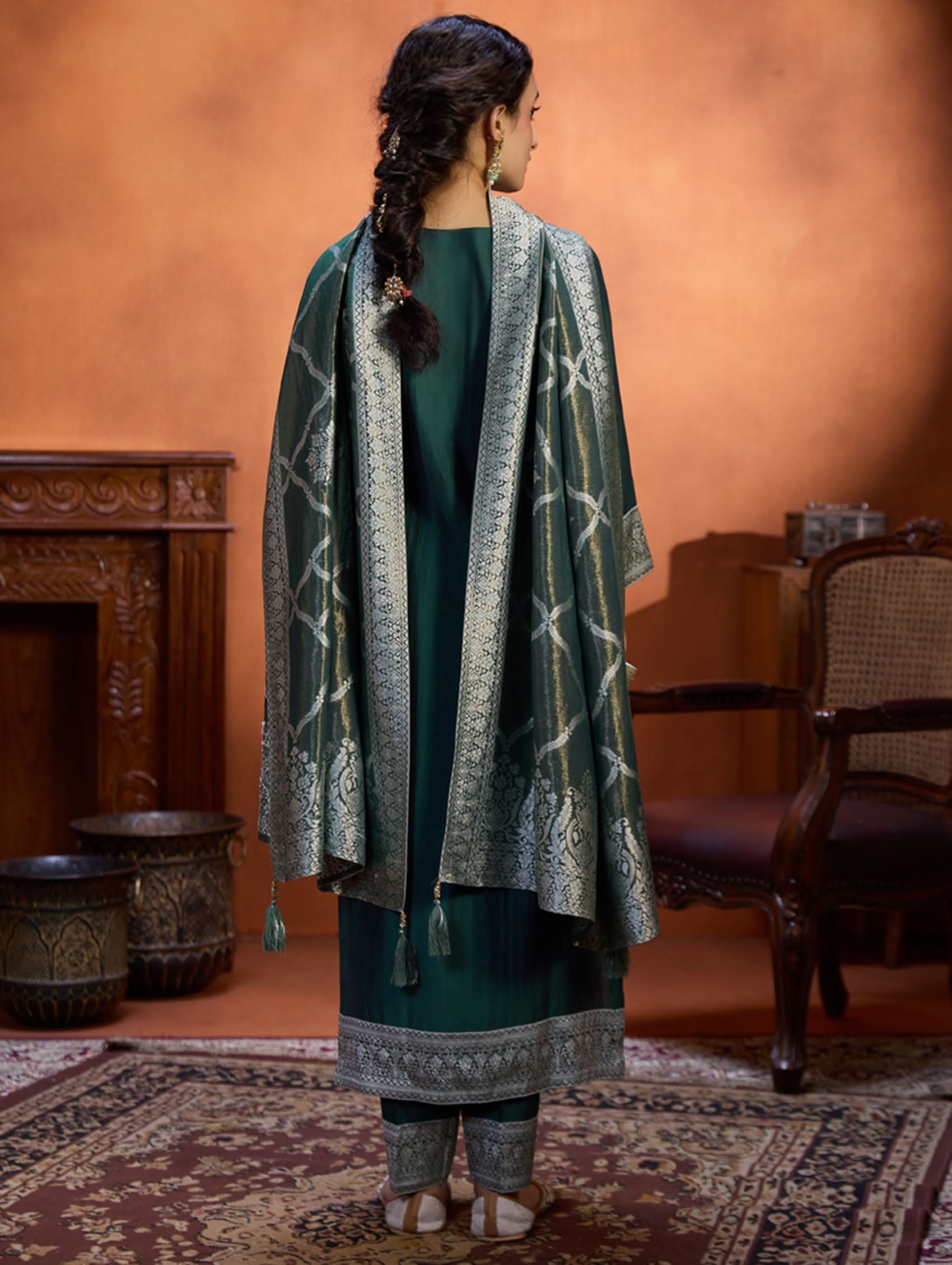 A woman wearing a green viscose silk suit set with a solid-coloured kurta, pants, and a jacquard weave shimmer silk dupatta, standing in a room with wooden furniture.