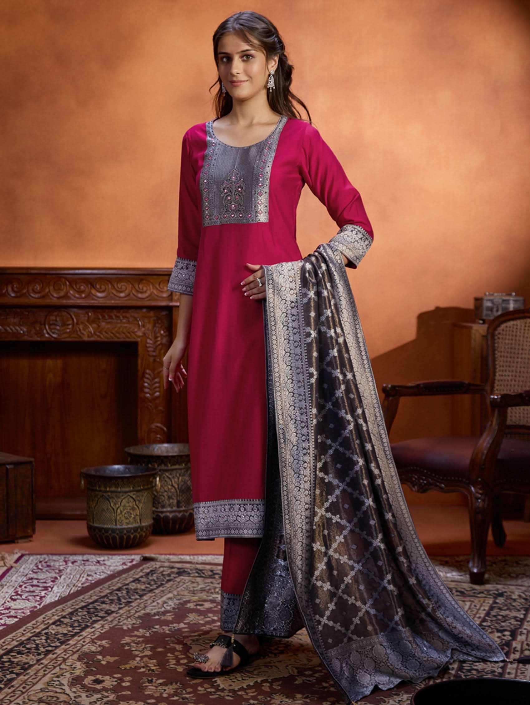 A woman wearing a traditional embroidered viscose silk suit set with a kurta, pants, and a dupatta in a room with an orange backdrop. The set is predominantly pink with embroidery details.