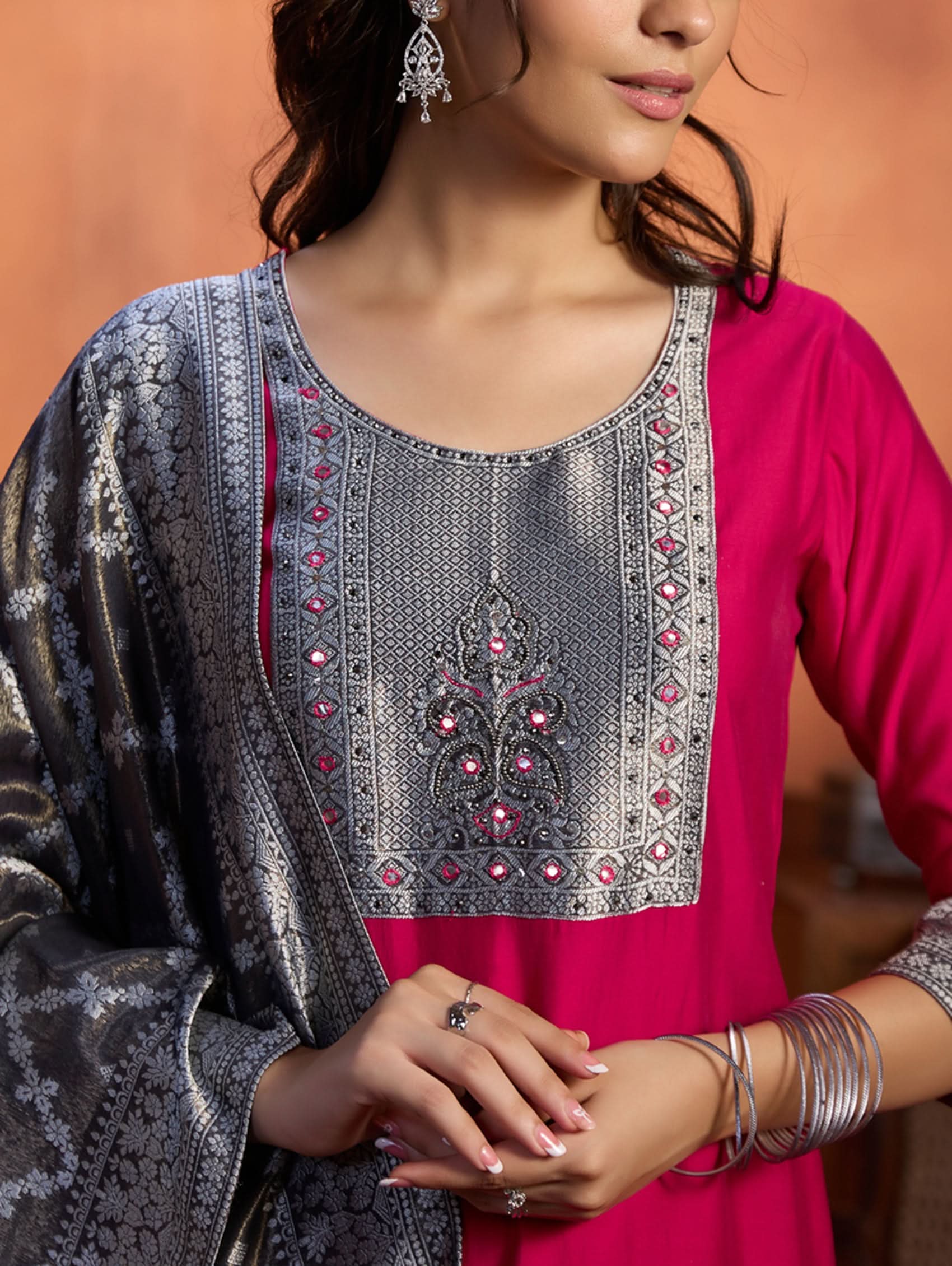 A woman wearing a traditional embroidered viscose silk suit set with a kurta, pants, and a dupatta in a room with an orange backdrop. The set is predominantly pink with embroidery details.