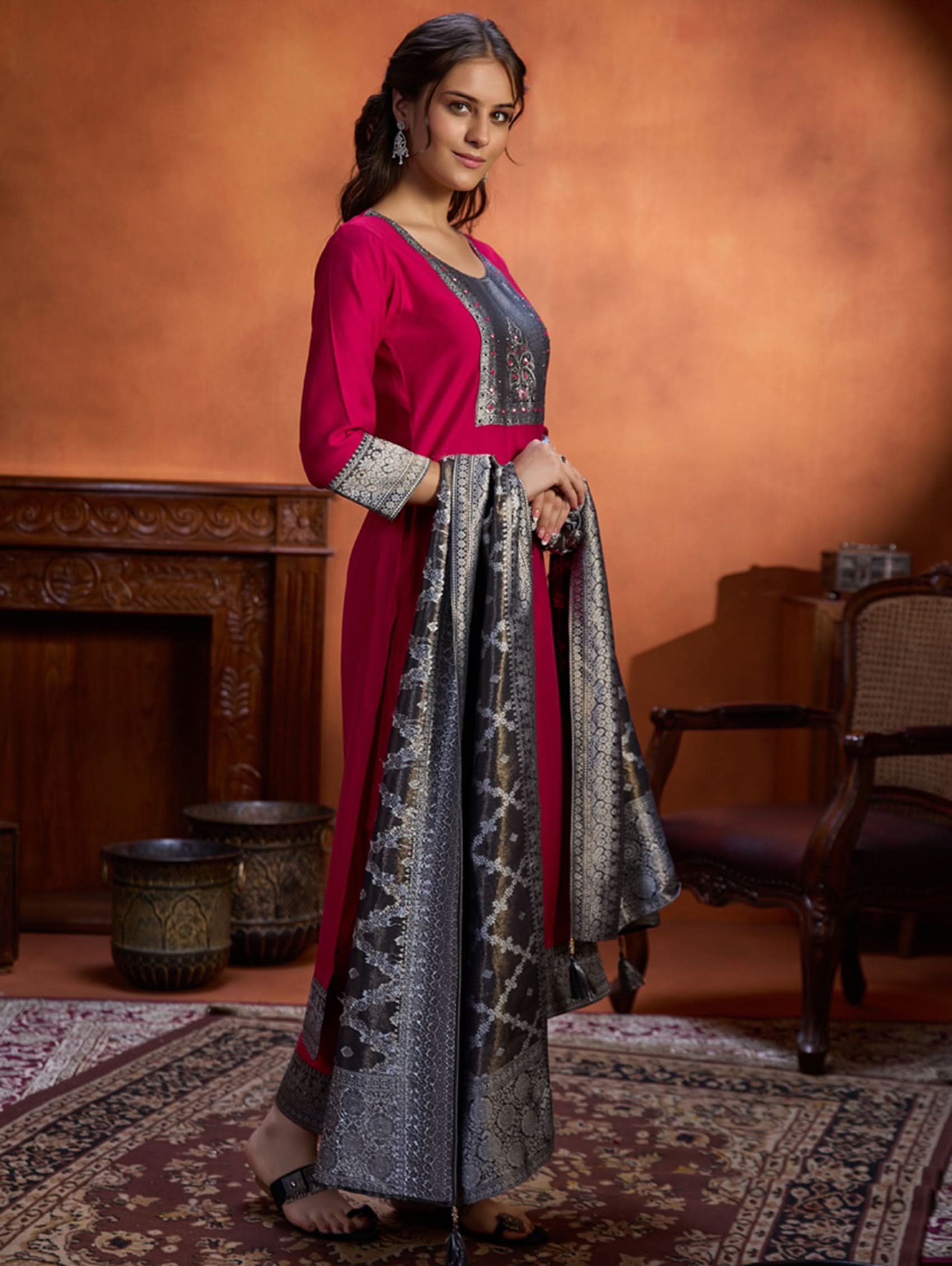 A woman wearing a traditional embroidered viscose silk suit set with a kurta, pants, and a dupatta in a room with an orange backdrop. The set is predominantly pink with embroidery details.