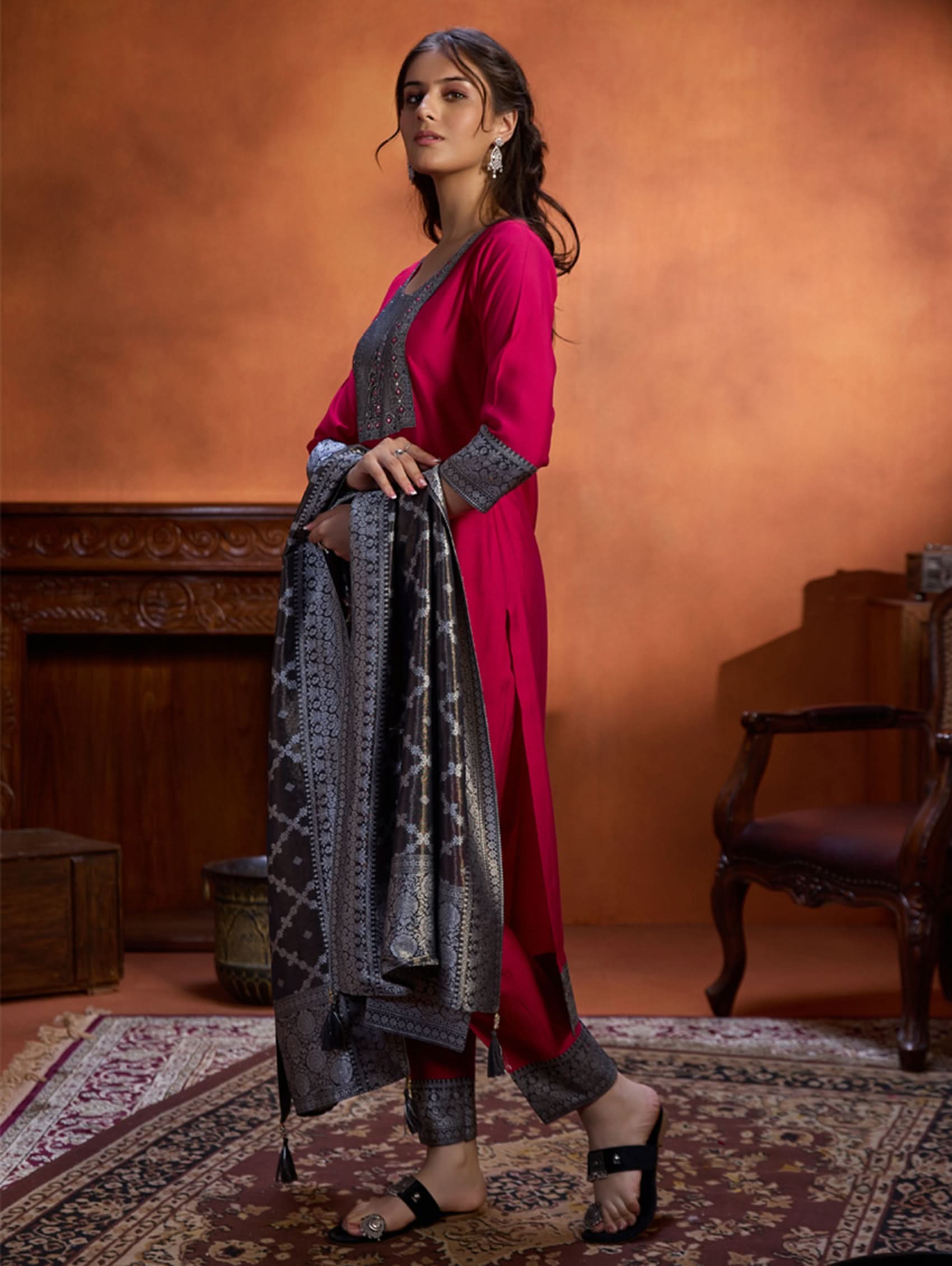 A woman wearing a traditional embroidered viscose silk suit set with a kurta, pants, and a dupatta in a room with an orange backdrop. The set is predominantly pink with embroidery details.