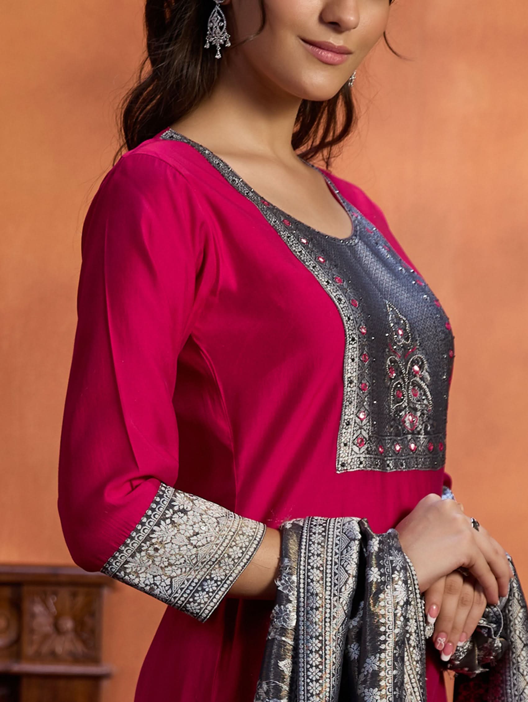 A woman wearing a traditional embroidered viscose silk suit set with a kurta, pants, and a dupatta in a room with an orange backdrop. The set is predominantly pink with embroidery details.