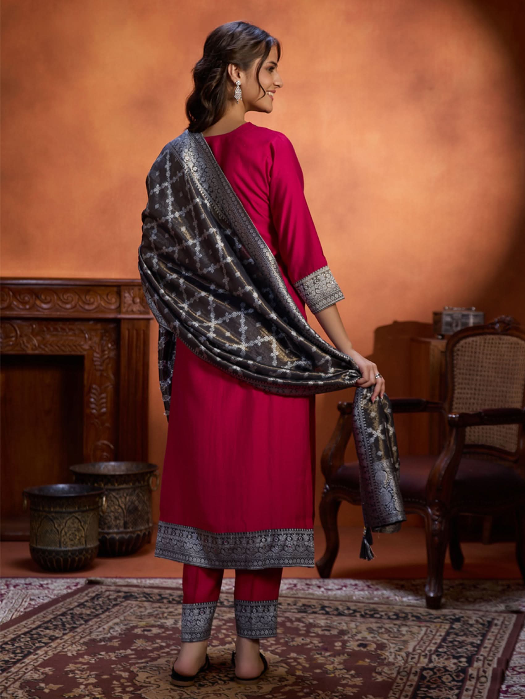 A woman wearing a traditional embroidered viscose silk suit set with a kurta, pants, and a dupatta in a room with an orange backdrop. The set is predominantly pink with embroidery details.