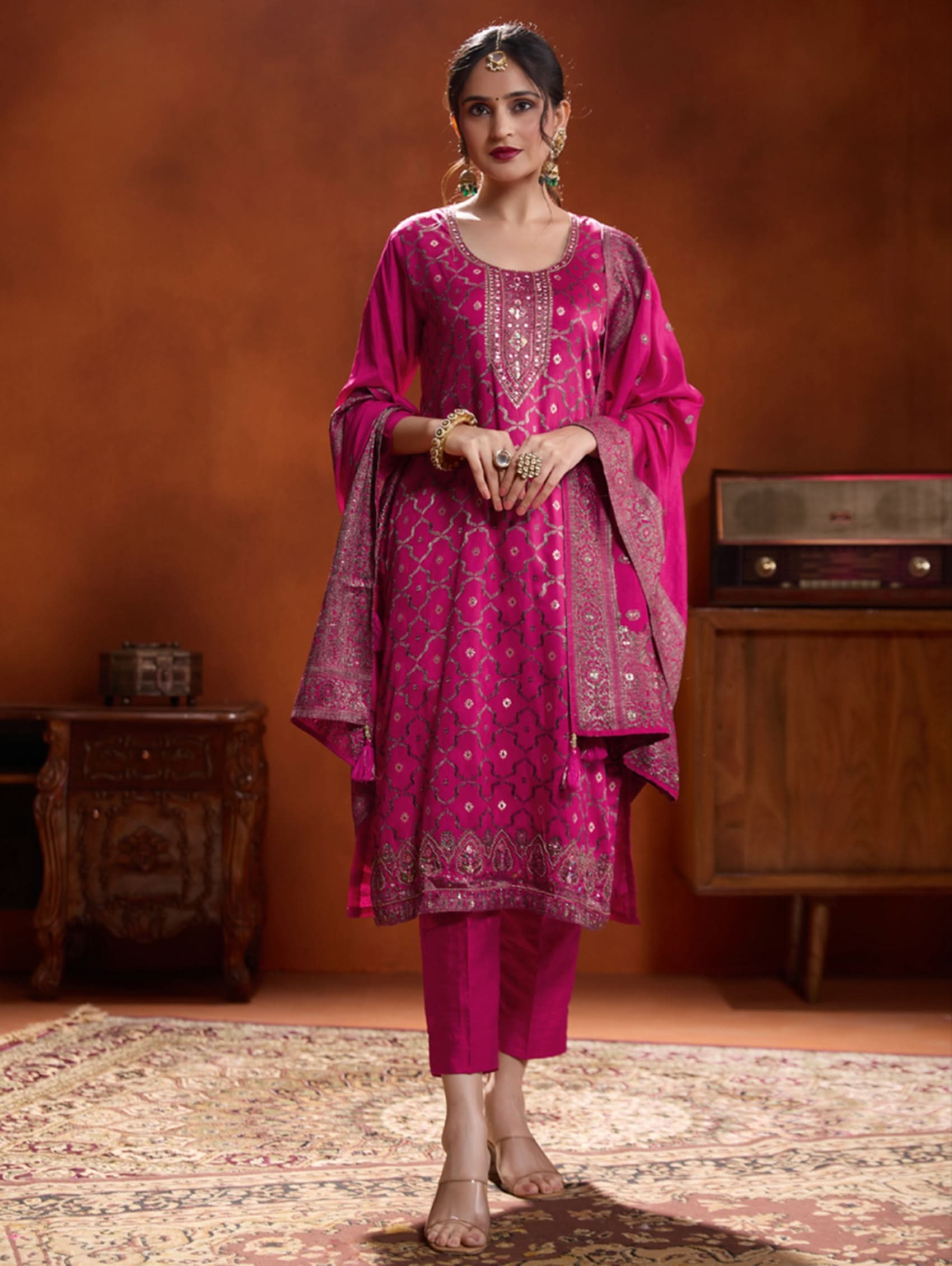 A woman wearing a pink jacquard weave silk suit set with hand-embroidered details, consisting of a kurta with three-fourth sleeves, a round neckline, and side slits, paired with pants and a dupatta.