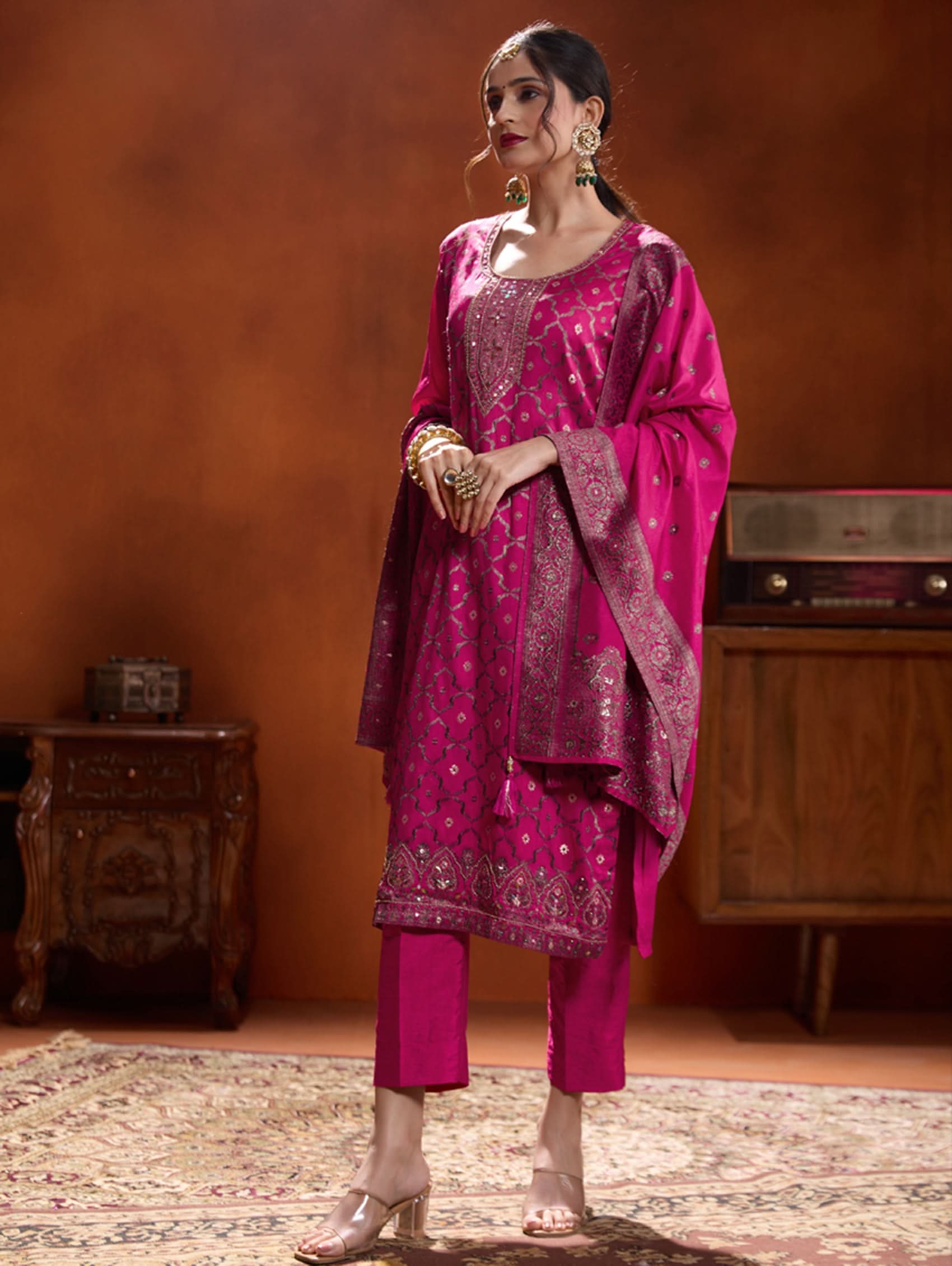 A woman wearing a pink jacquard weave silk suit set with hand-embroidered details, consisting of a kurta with three-fourth sleeves, a round neckline, and side slits, paired with pants and a dupatta.