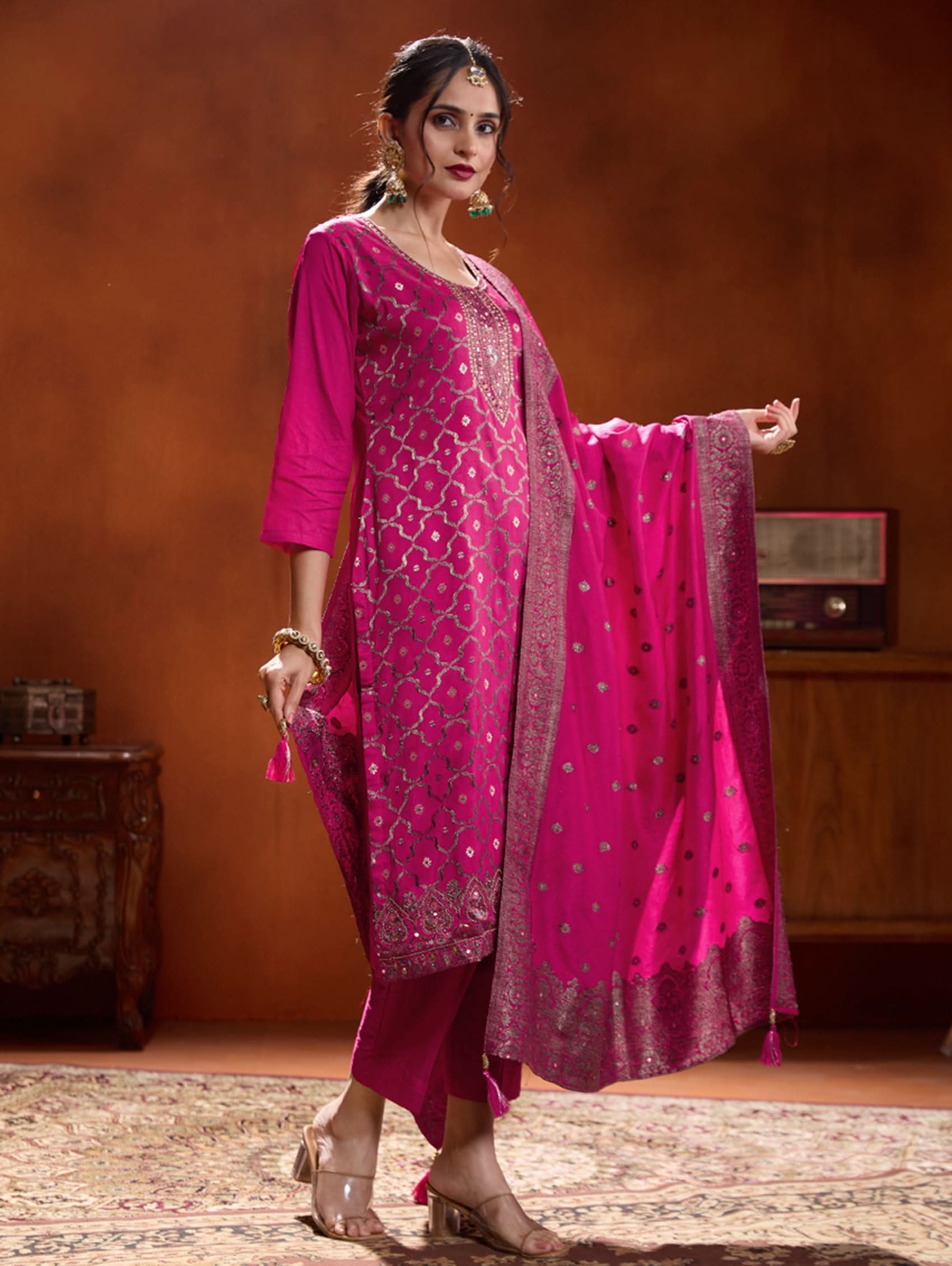 A woman wearing a pink jacquard weave silk suit set with hand-embroidered details, consisting of a kurta with three-fourth sleeves, a round neckline, and side slits, paired with pants and a dupatta.