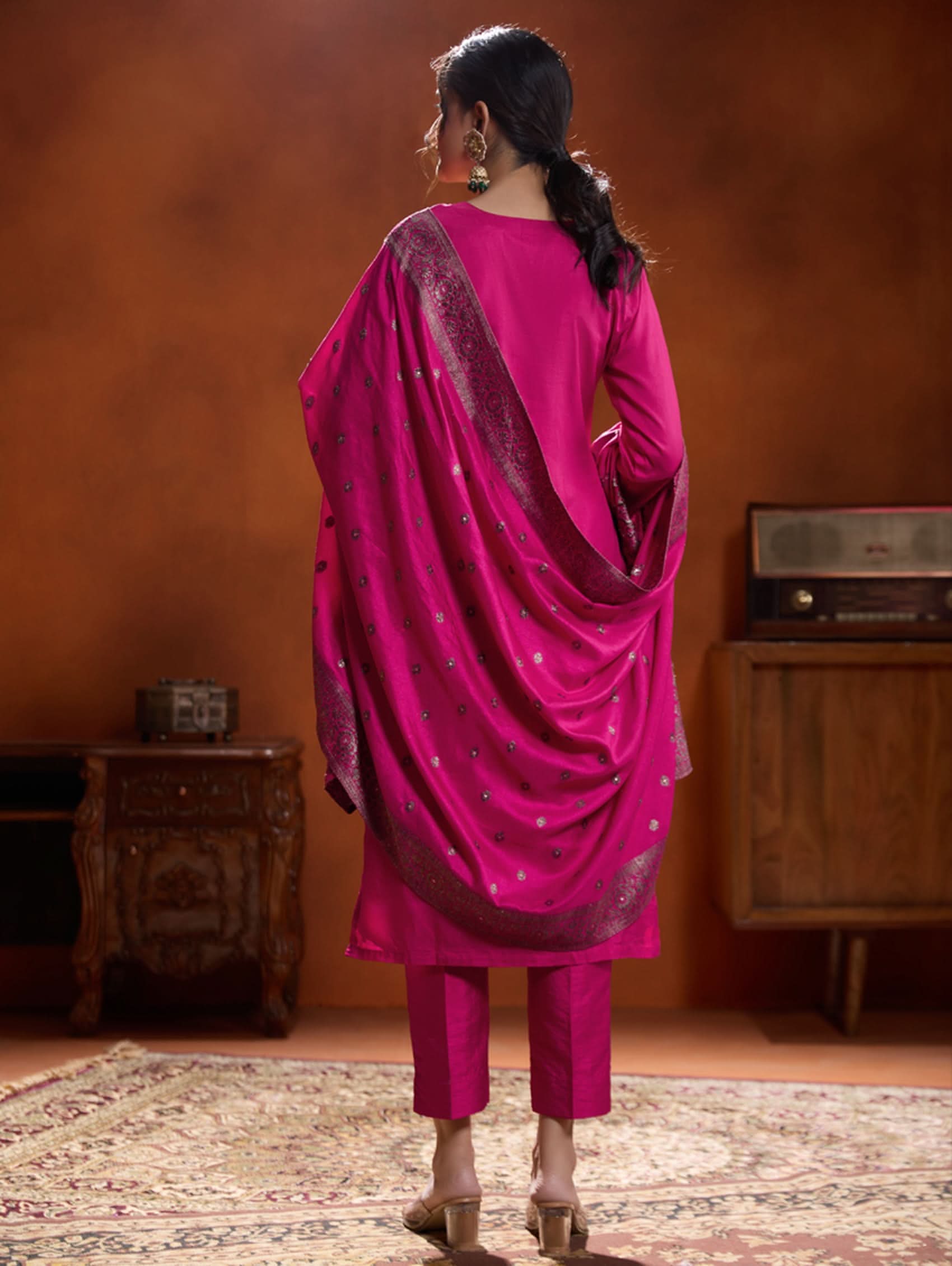 A woman wearing a pink jacquard weave silk suit set with hand-embroidered details, consisting of a kurta with three-fourth sleeves, a round neckline, and side slits, paired with pants and a dupatta.