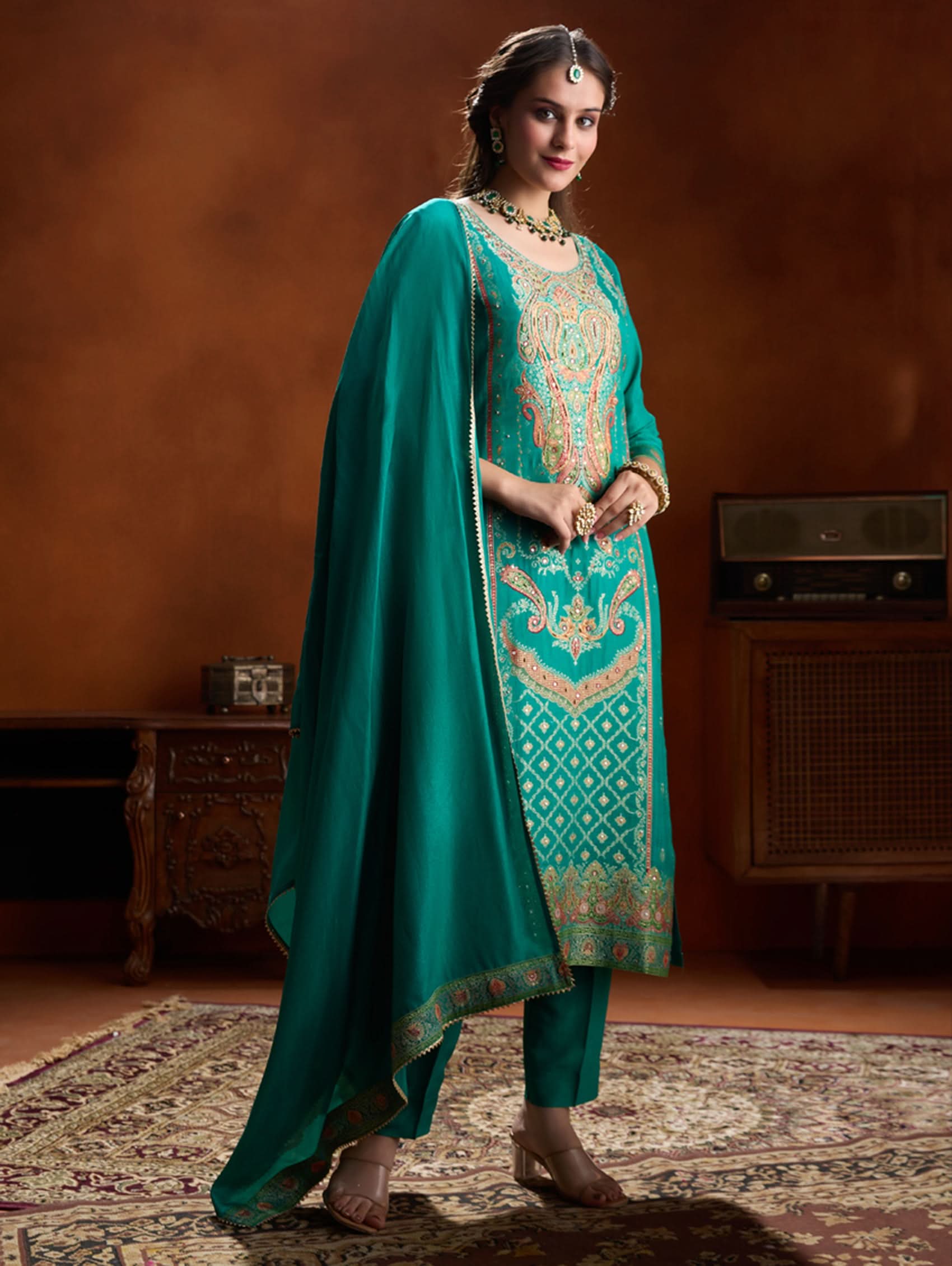 Woman in a teal embroidered outfit standing in a room with a warm brown wall.