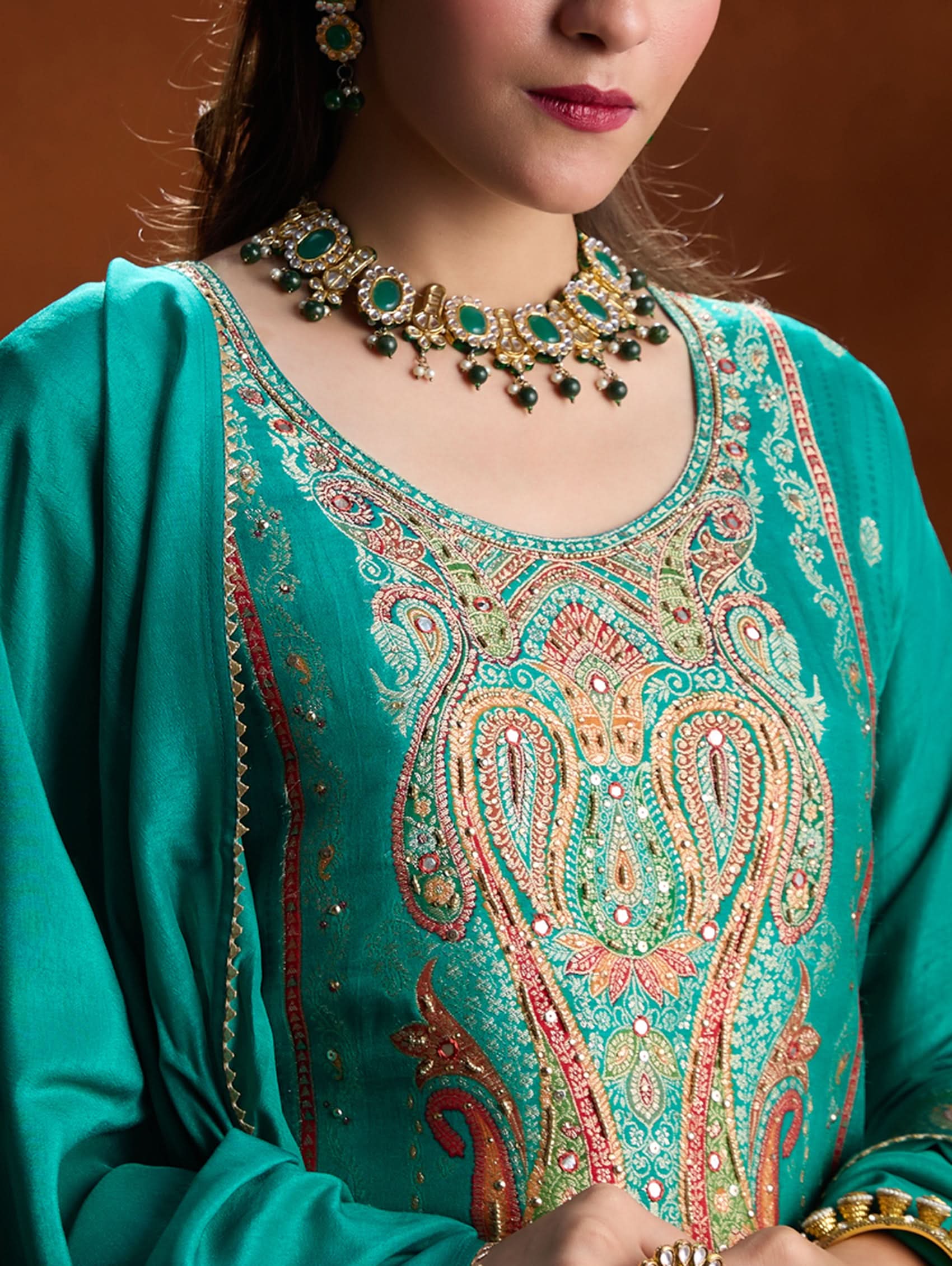 Teal embroidered garment with intricate patterns and jewelry on a brown background